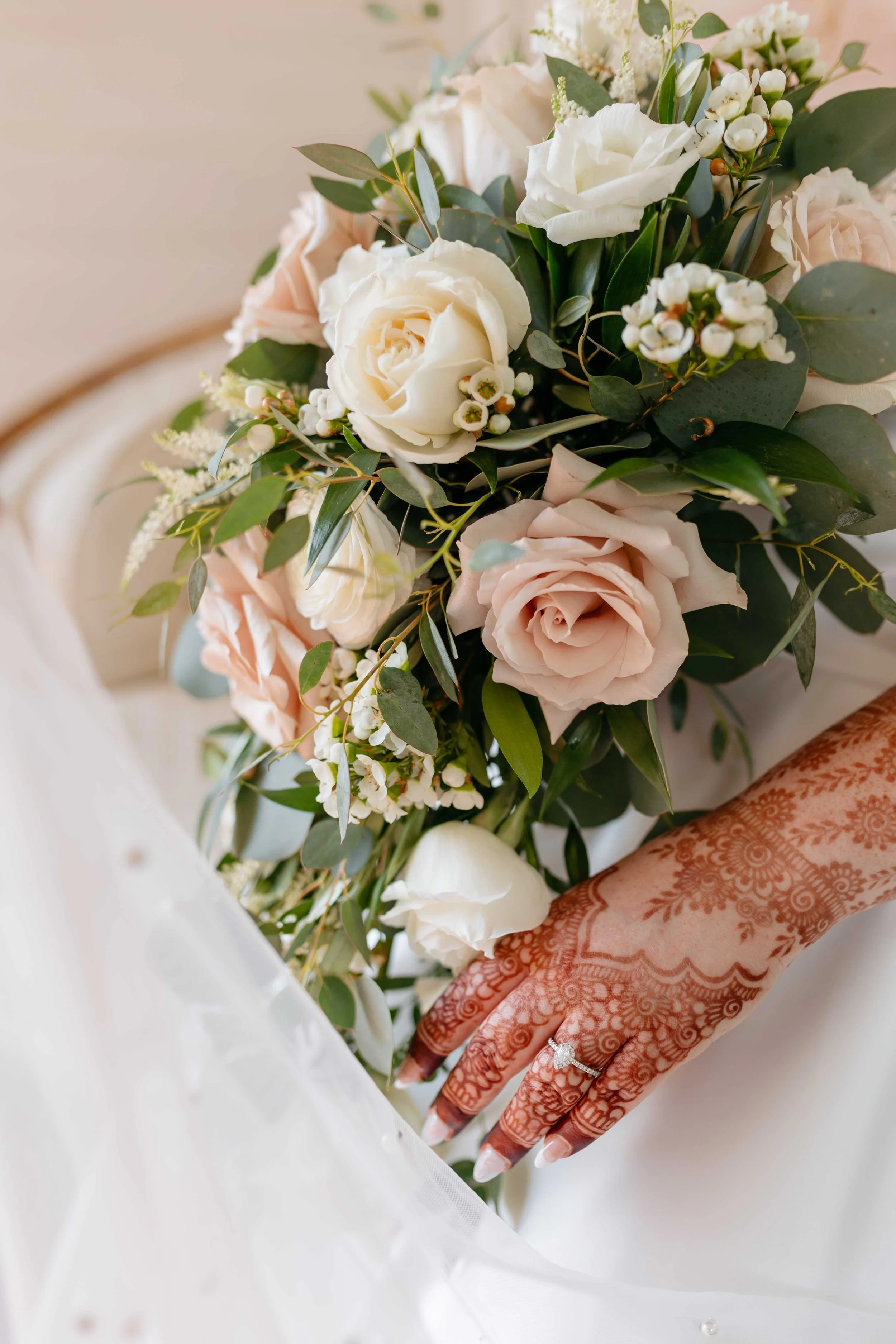 Close-up of a hand with intricate henna designs and a wedding ring, holding a bouquet of white and pale pink roses, white small flowers, and greenery.
