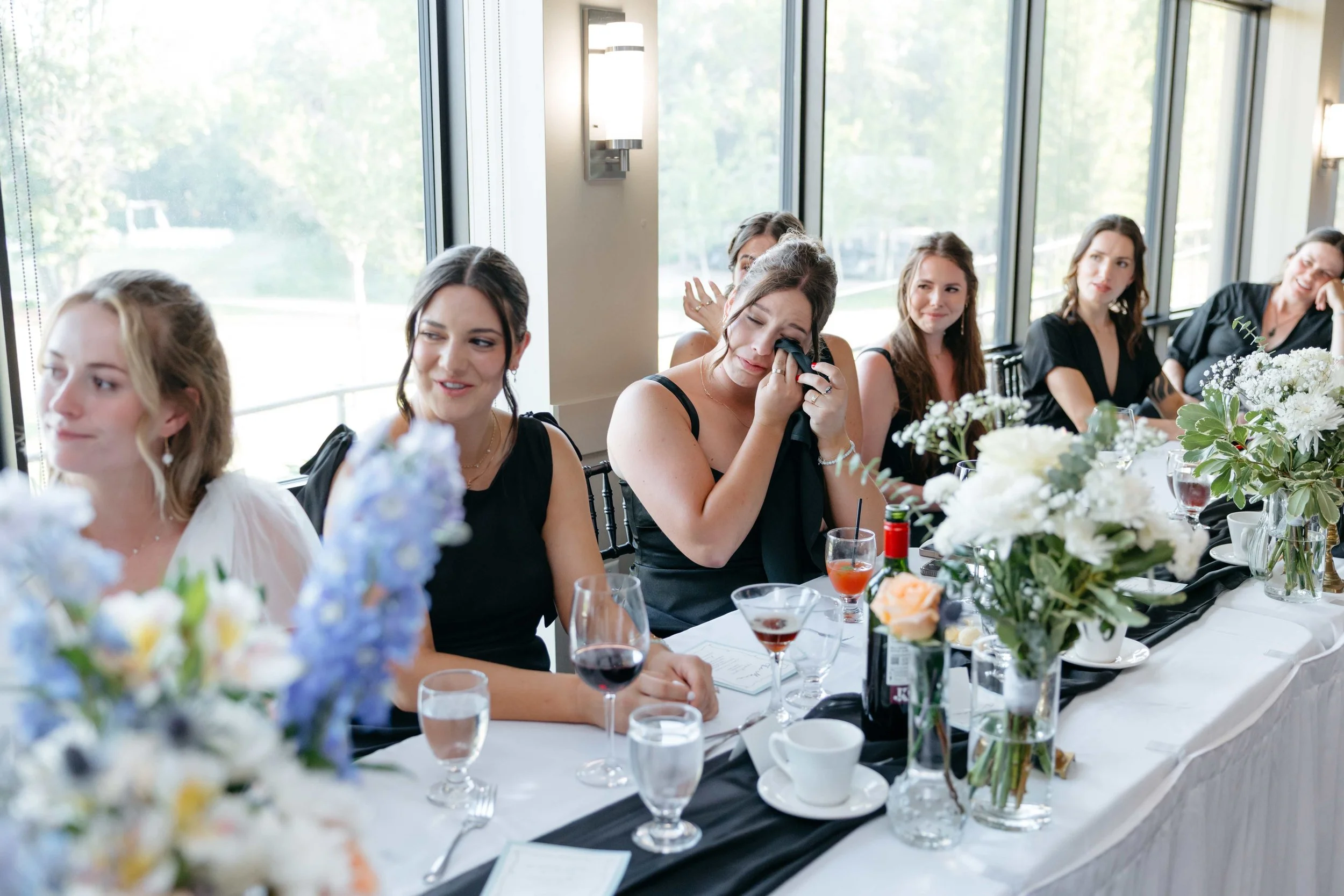 A group of women sitting at a long table during a celebration or wedding reception, with various flower arrangements and drinks on the table, near large windows letting in natural light.