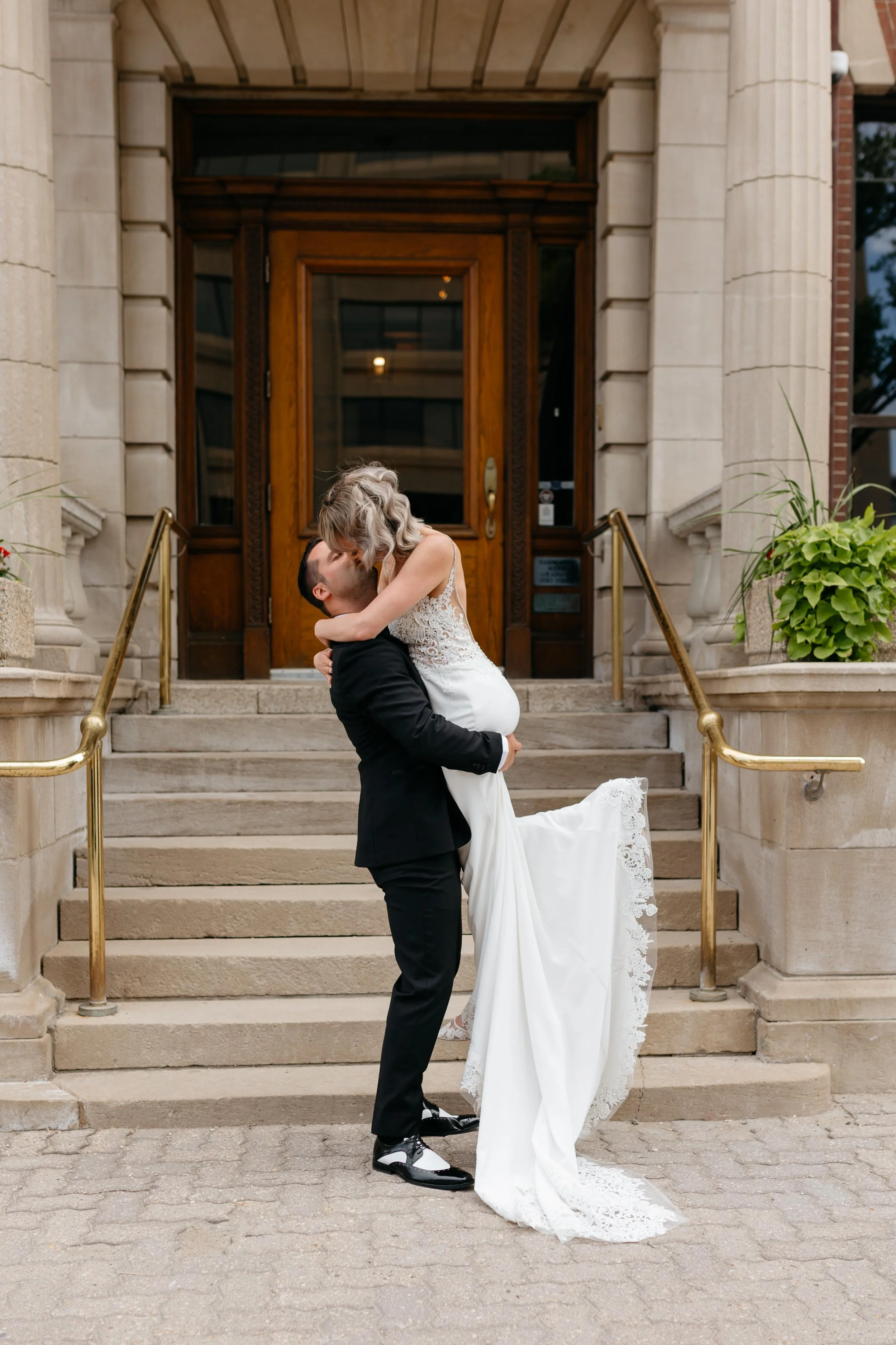 A newlywed couple sharing a kiss in front of a grand stone building with stairs and brass handrails. The groom is lifting the bride, who is wearing a white wedding dress with lace details, while the groom wears a black suit and sneakers.