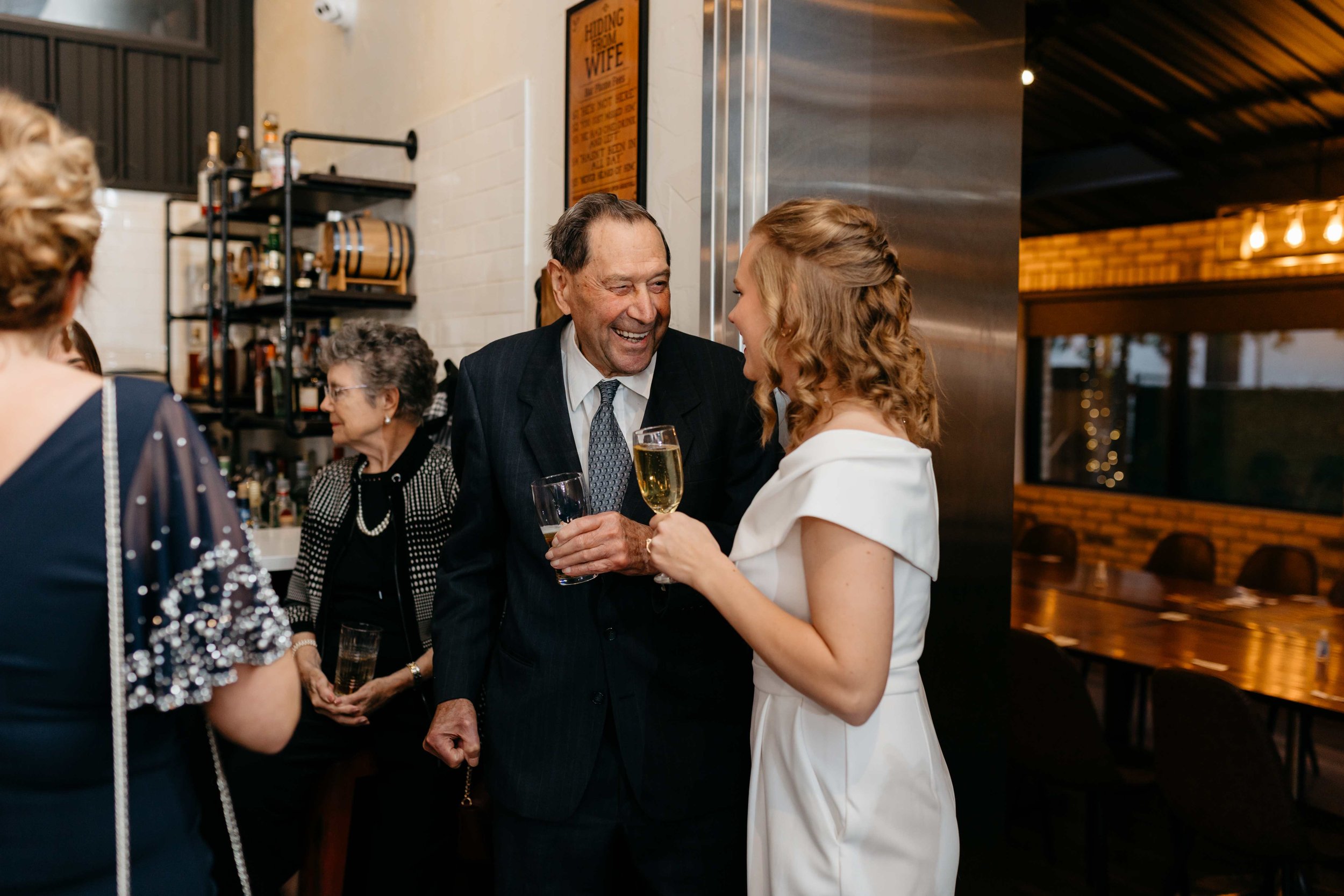 A group of people at a social event, with a man in a dark suit and a woman in a white dress smiling and holding champagne glasses, engaging in conversation.