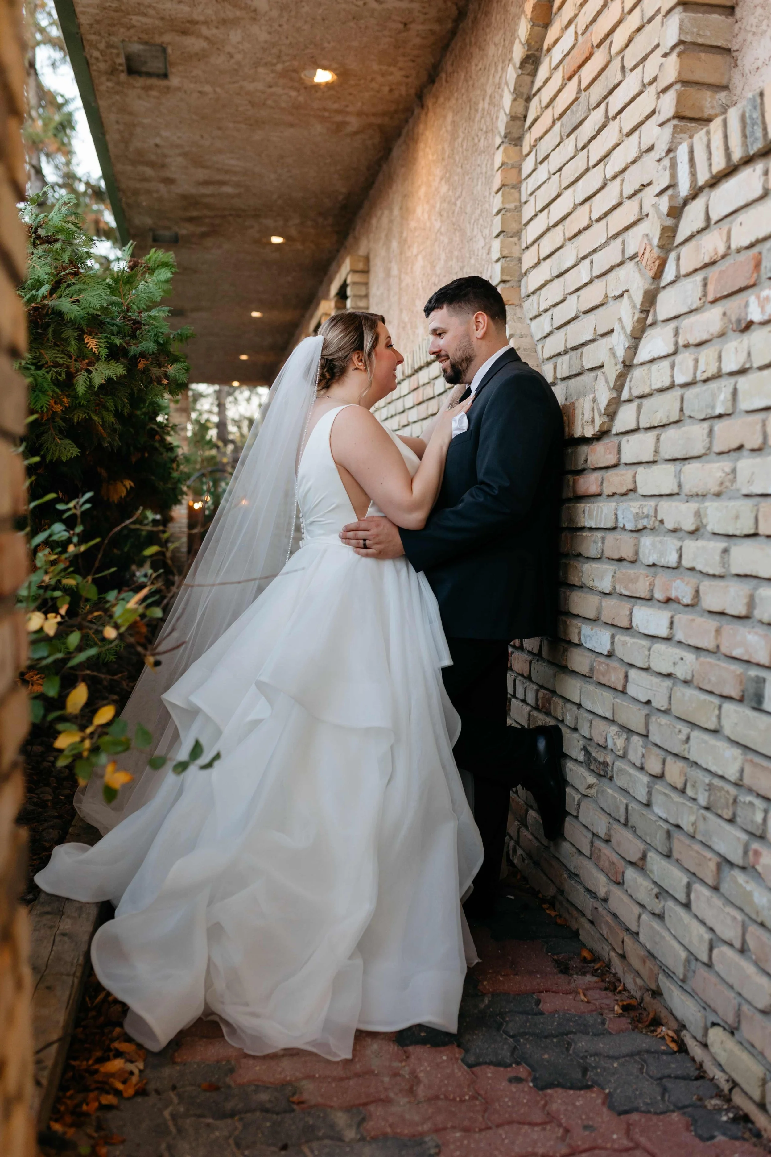 A bride and groom standing close, facing each other with smiles. The bride is wearing a white wedding gown and veil, and the groom is in a black suit with a white shirt. They are outdoors against a brick wall, with greenery on the left and a brick pa
