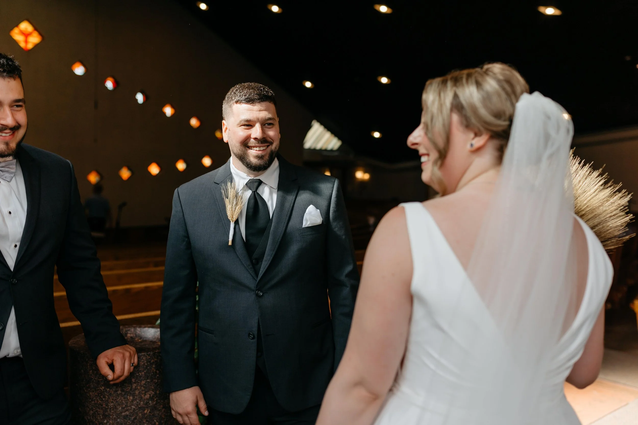 A group of people dressed formally at a wedding, with a woman in a white dress and veil smiling and talking to a man in a tuxedo, with two other men also in tuxedos nearby, in a decorated indoor venue.