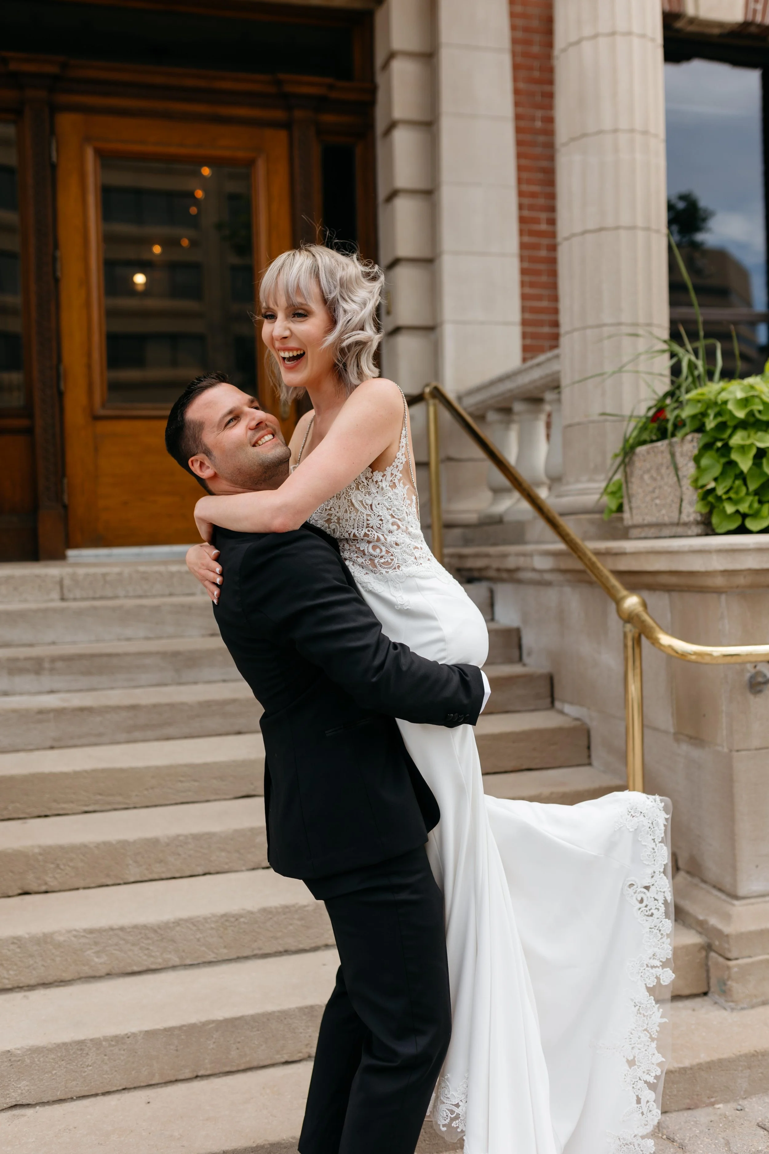A happy bride in a white wedding gown with lace details being lifted by a smiling groom in a black suit in front of a building with steps and columns.