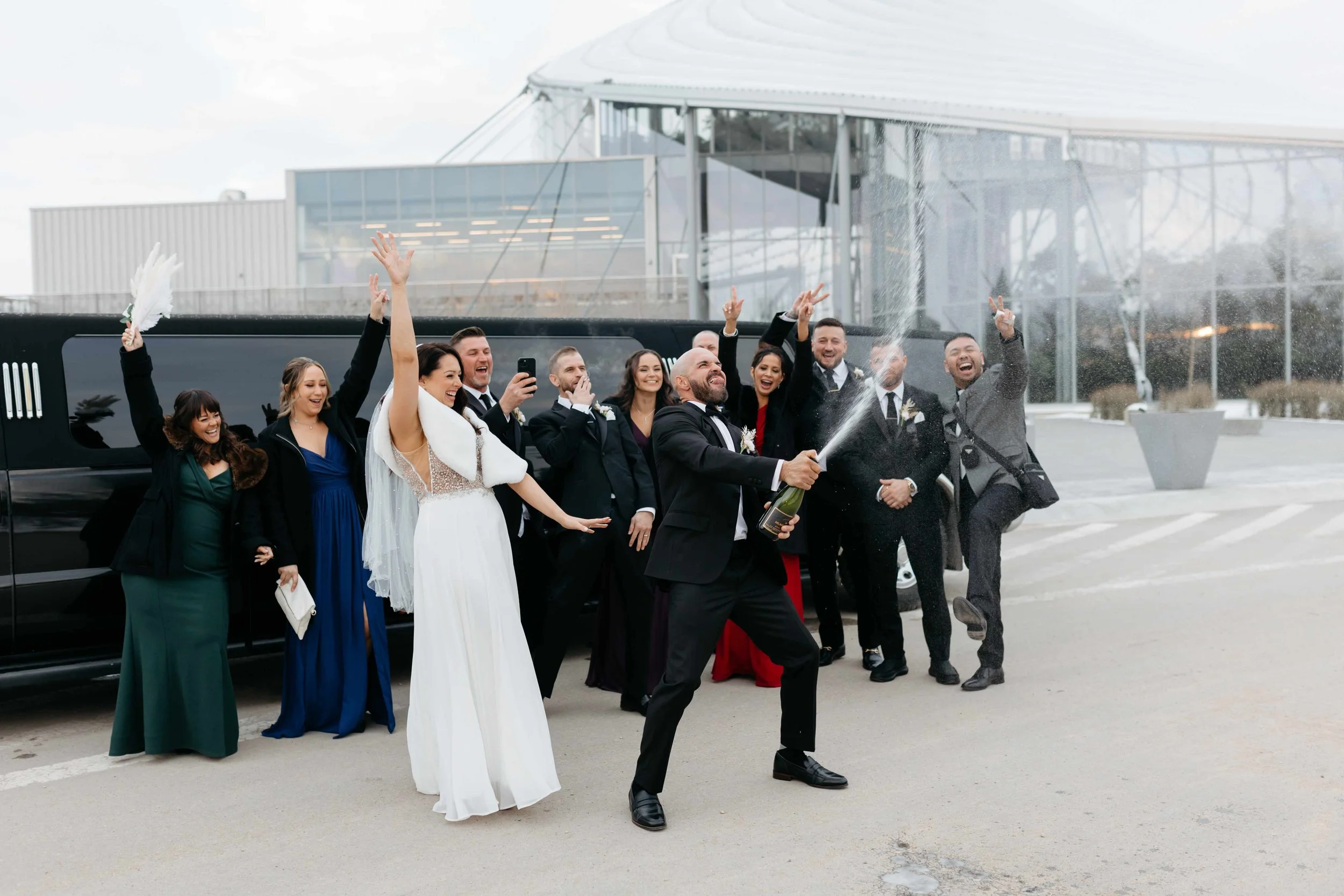Group of people in formal attire celebrating, some spraying champagne, outside a modern building.