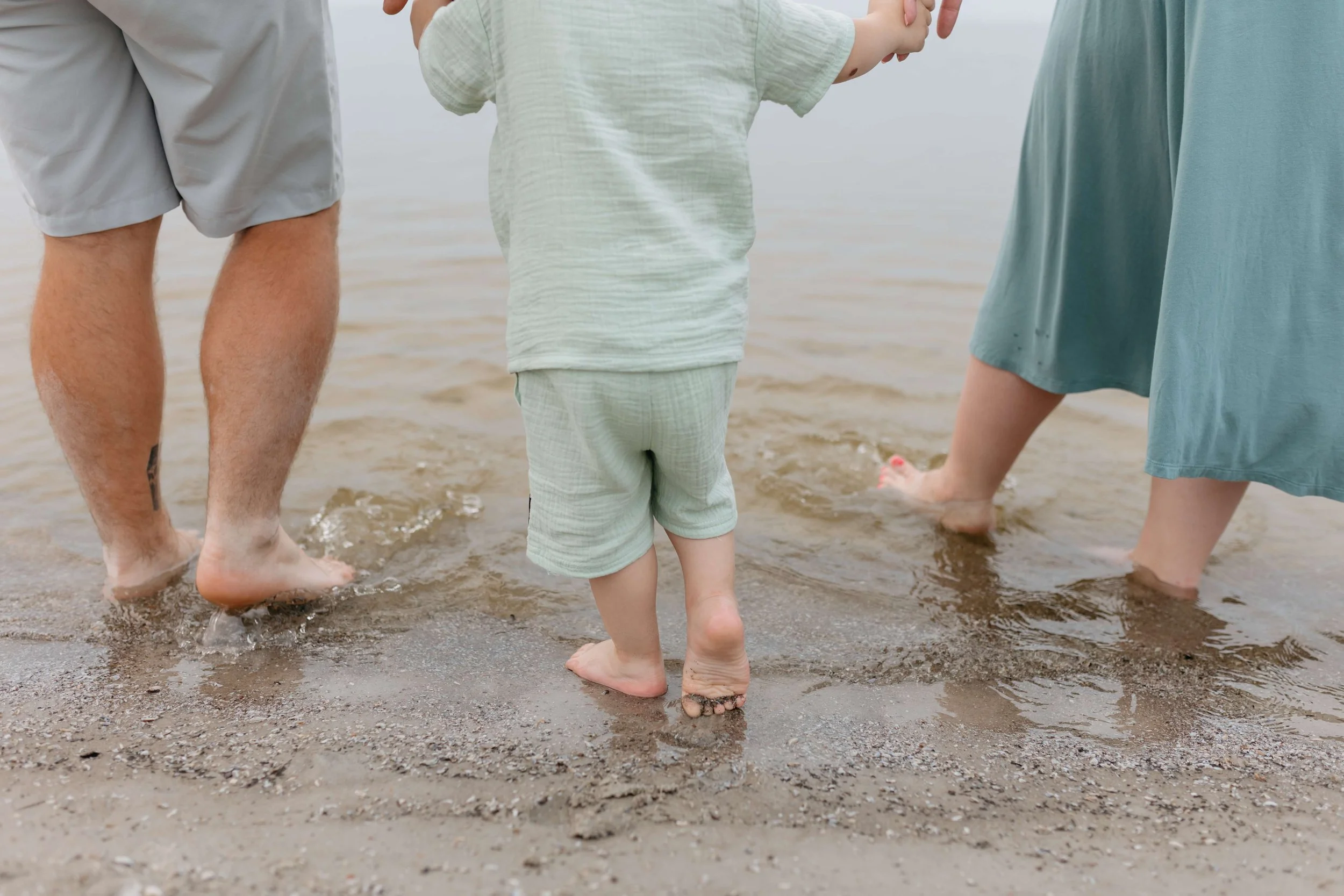 Parents and child, stand in the shallow water at a sandy beach in winnipeg during family session with sari risbey