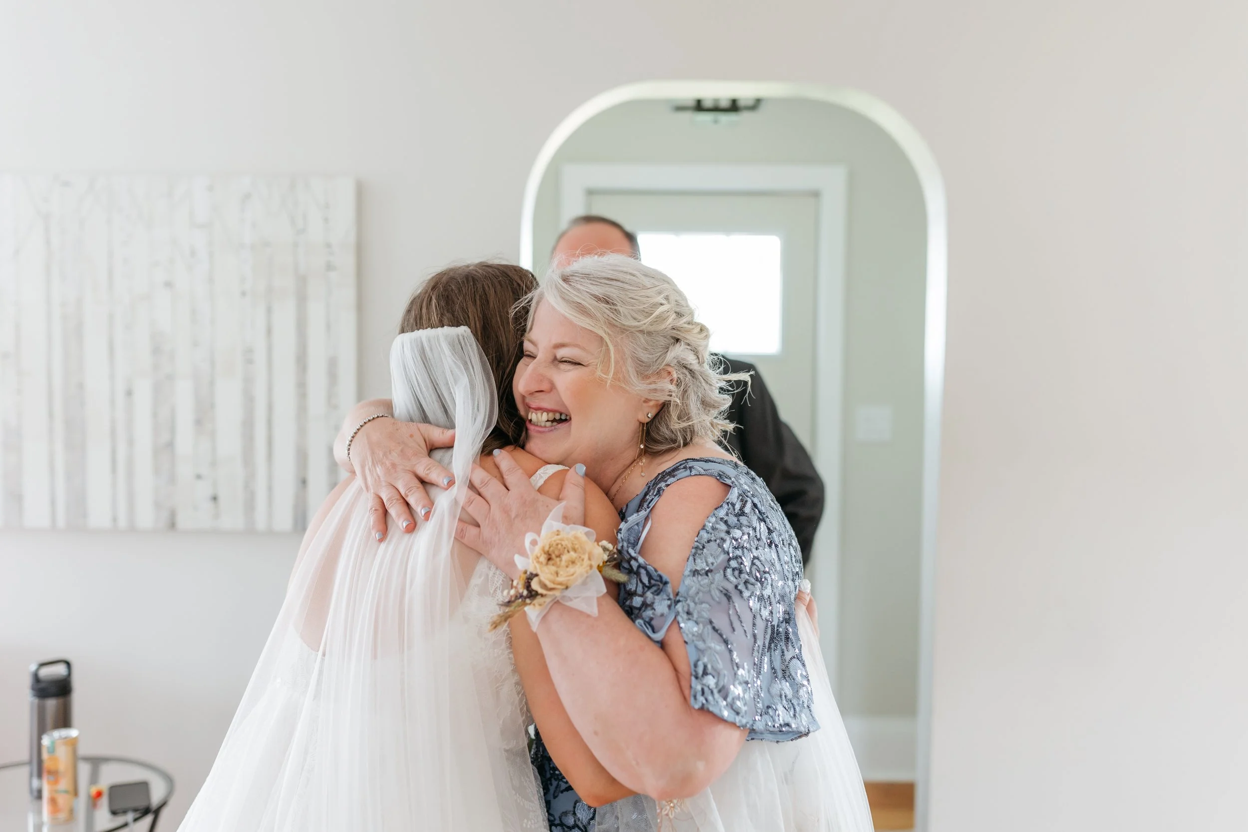 Mom hugging a bride in a wedding dress with a veil, both smiling, in a bright room with white walls and a mirror in the background. Sari Risbey Winnipeg wedding photographer.