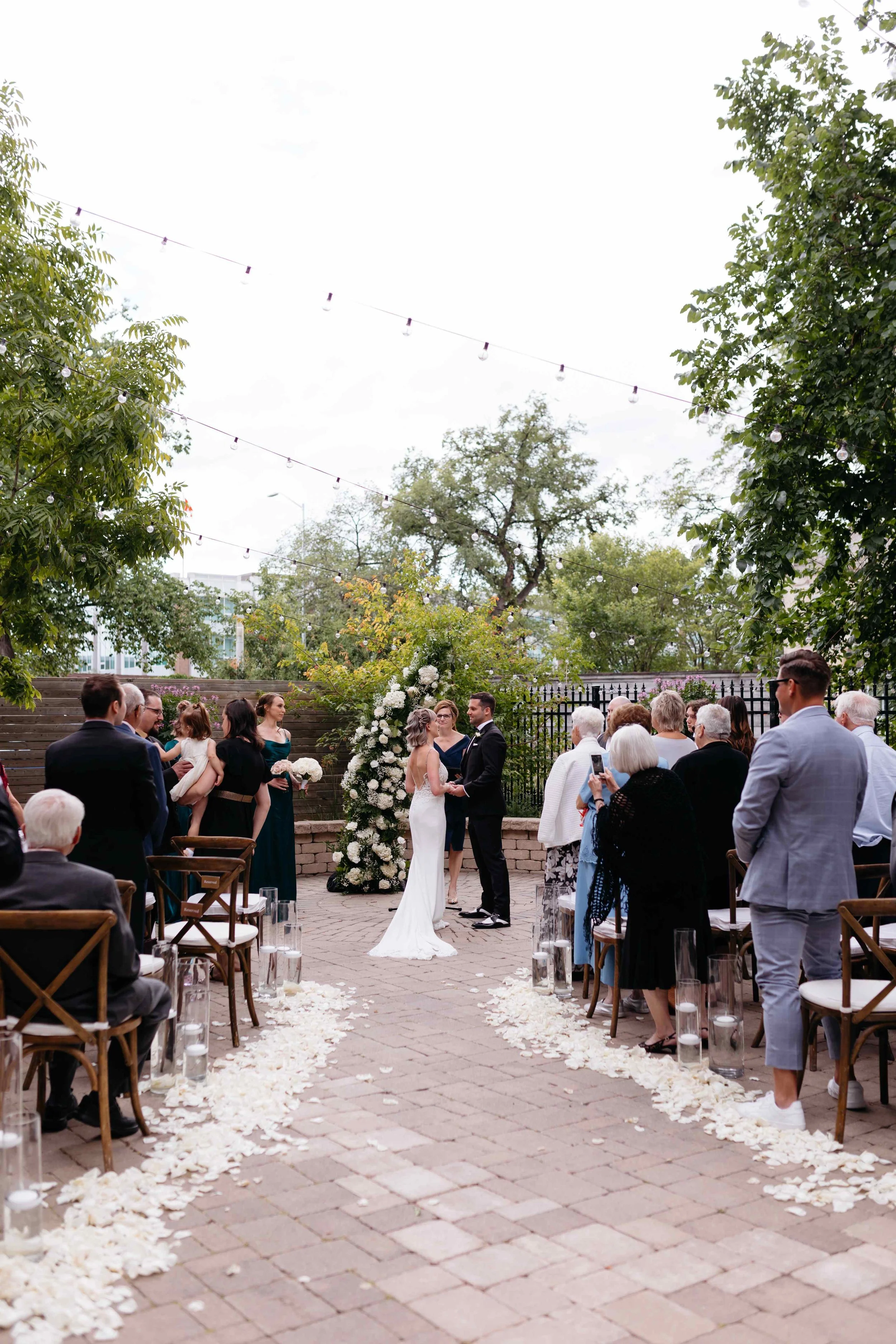 A wedding ceremony taking place outdoors with a bride and groom, surrounded by guests. The scene includes a floral arch and petals scattered along a brick pathway. Overhead string lights are visible in the sky, and trees frame the area.