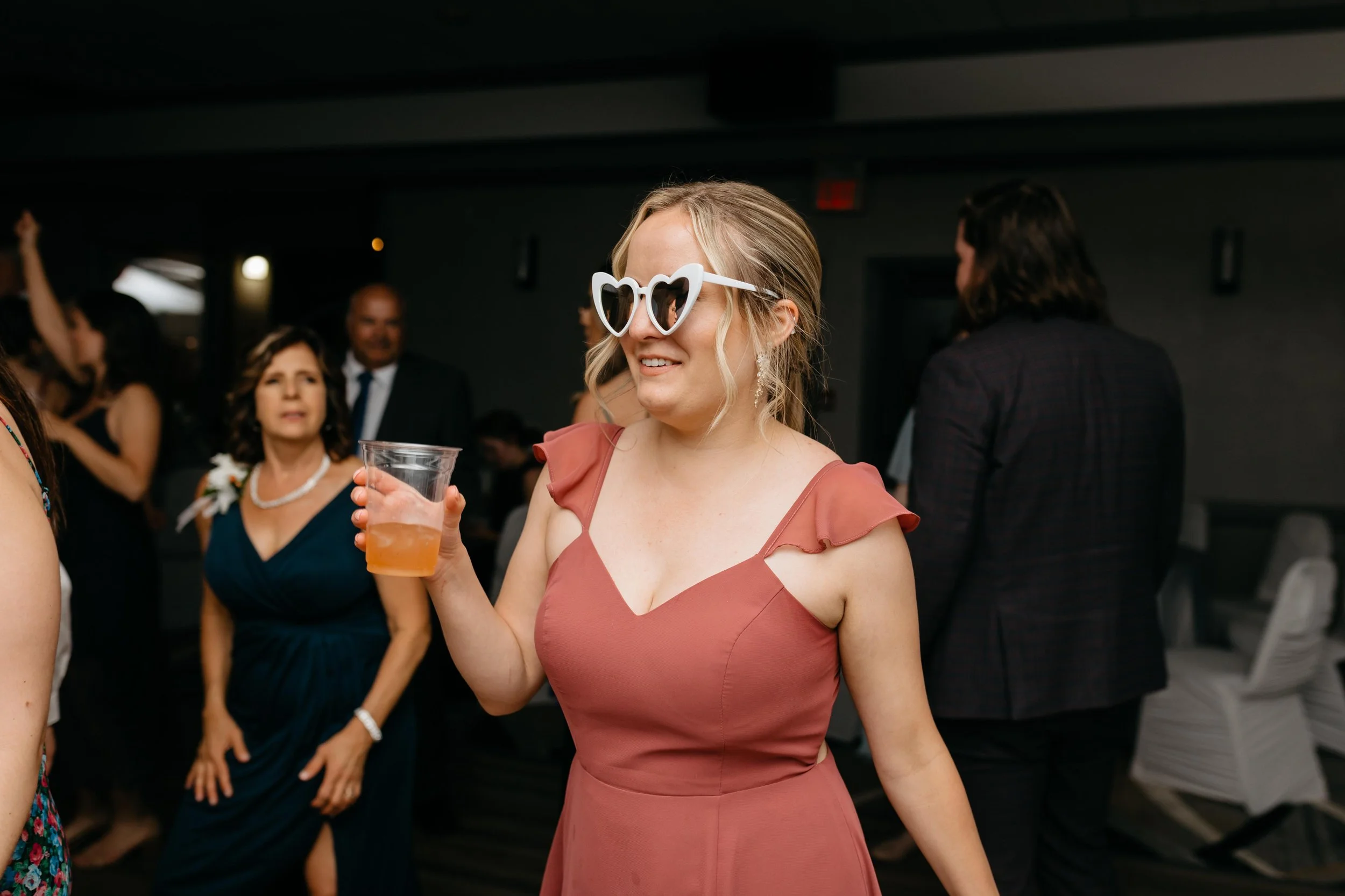 A woman at a social event wears heart-shaped sunglasses and holds a drink, smiling. Other guests are in the background, some talking and dressed formally.