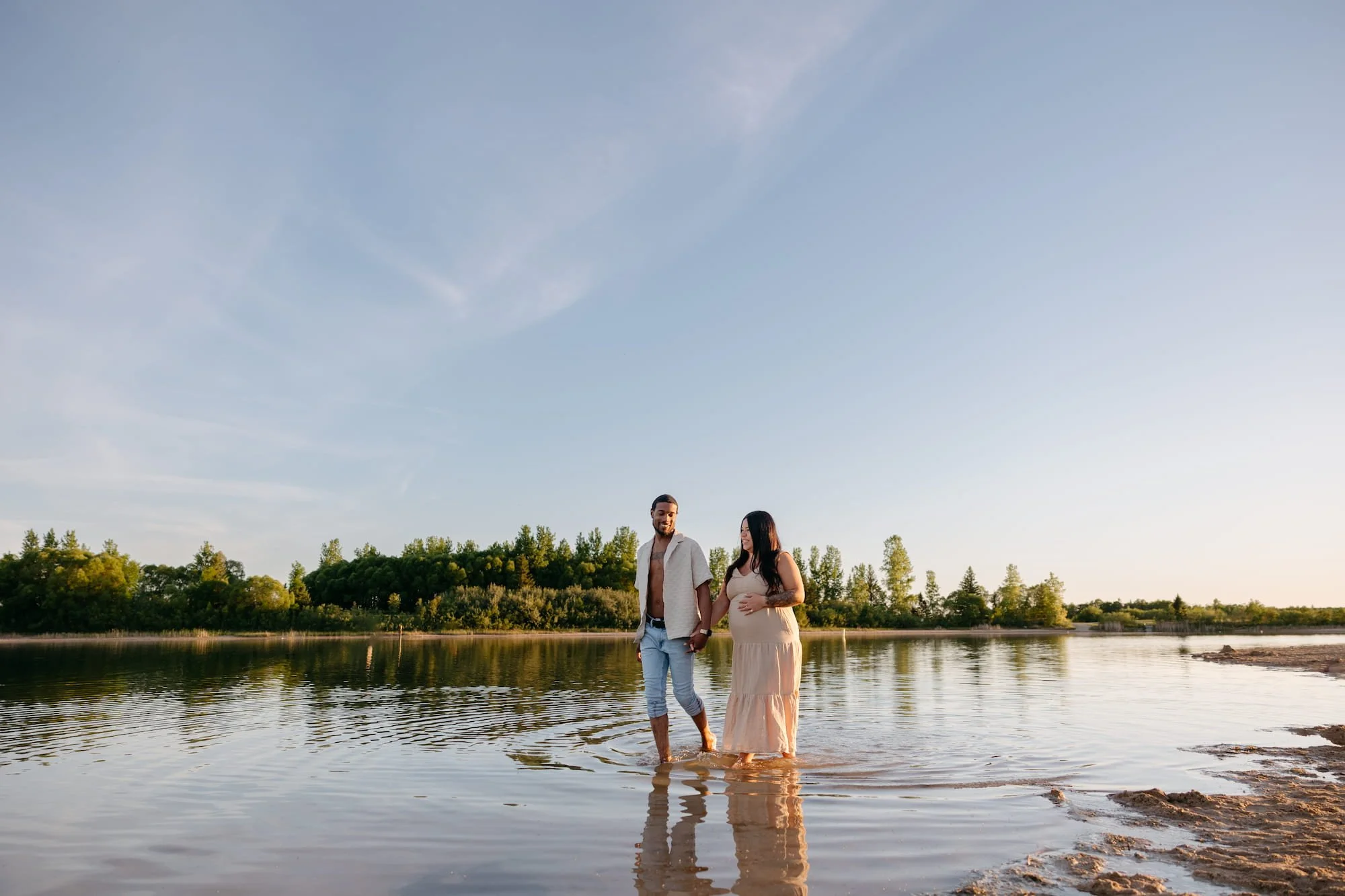 Pregnant couple walking hand in hand through the water during a relaxed maternity photography session in Winnipeg, Manitoba.