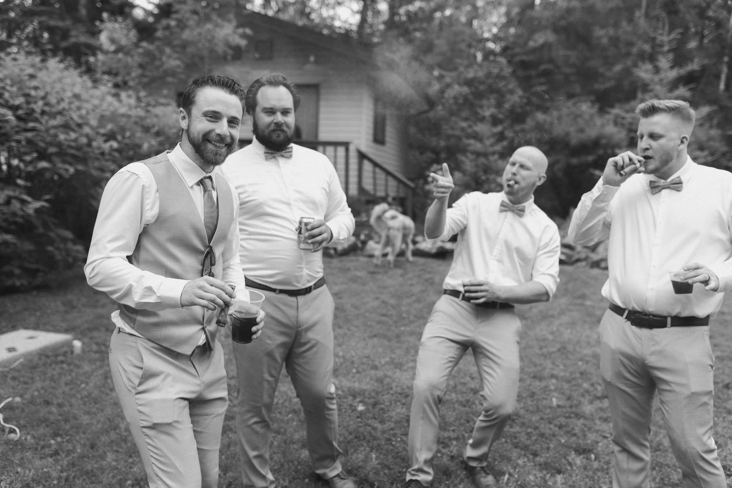 Four men at an outdoor gathering wearing formal attire, holding drinks, and socializing. One man is puffing on a cigarette or cigar, while others are smiling or gesturing. The background includes trees, a house, and a dog.