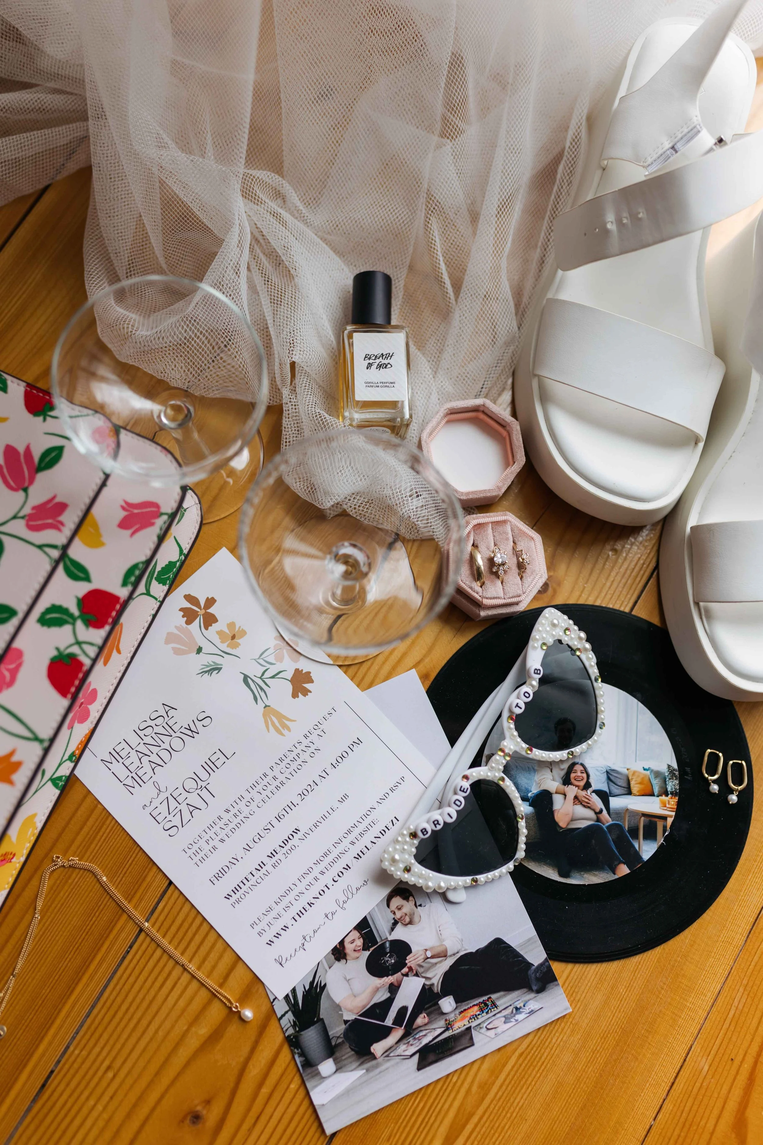 Decorative wedding invitation with floral design and information about the wedding reception. Items include glasses, perfume, jewelry, and accessories placed on a wooden surface with a draped fabric backdrop.