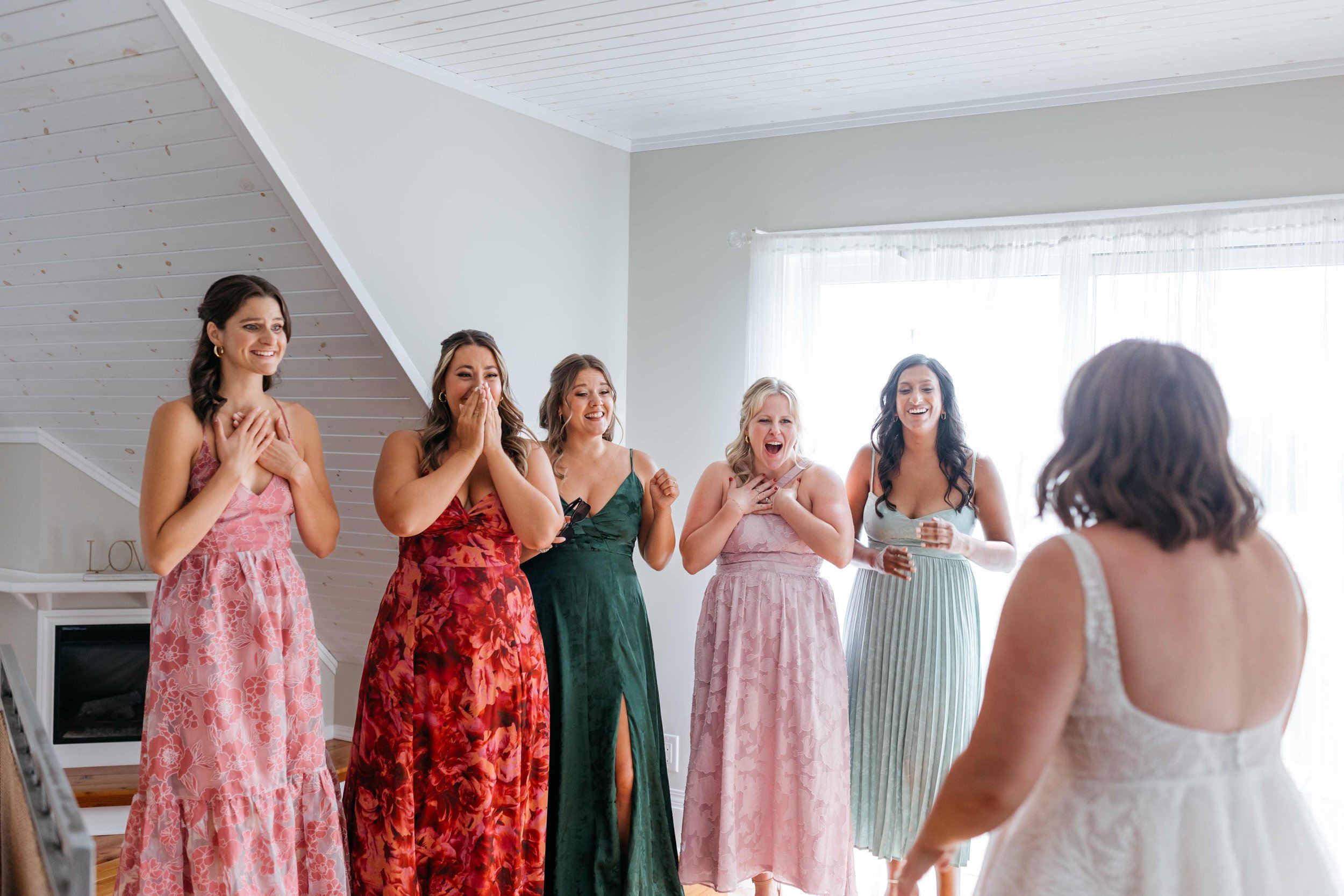 Women in colorful dresses are standing indoors, reacting with surprise and joy to a young girl in a white dress, likely during a special celebration or event.
