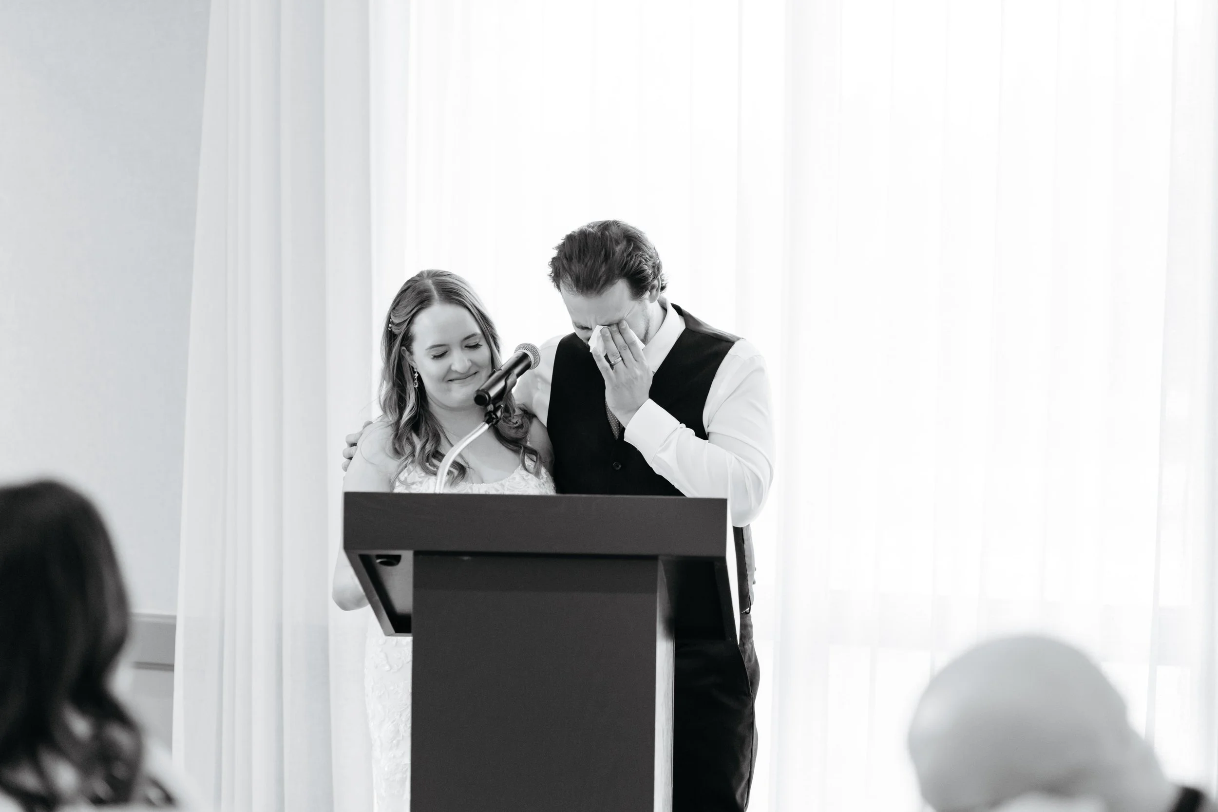 A man in a vest appears emotional, wiping his eye with a tissue, while a woman in a wedding dress smiles and places her hand on his shoulder, standing at a podium with a microphone, against a backdrop of white curtains.