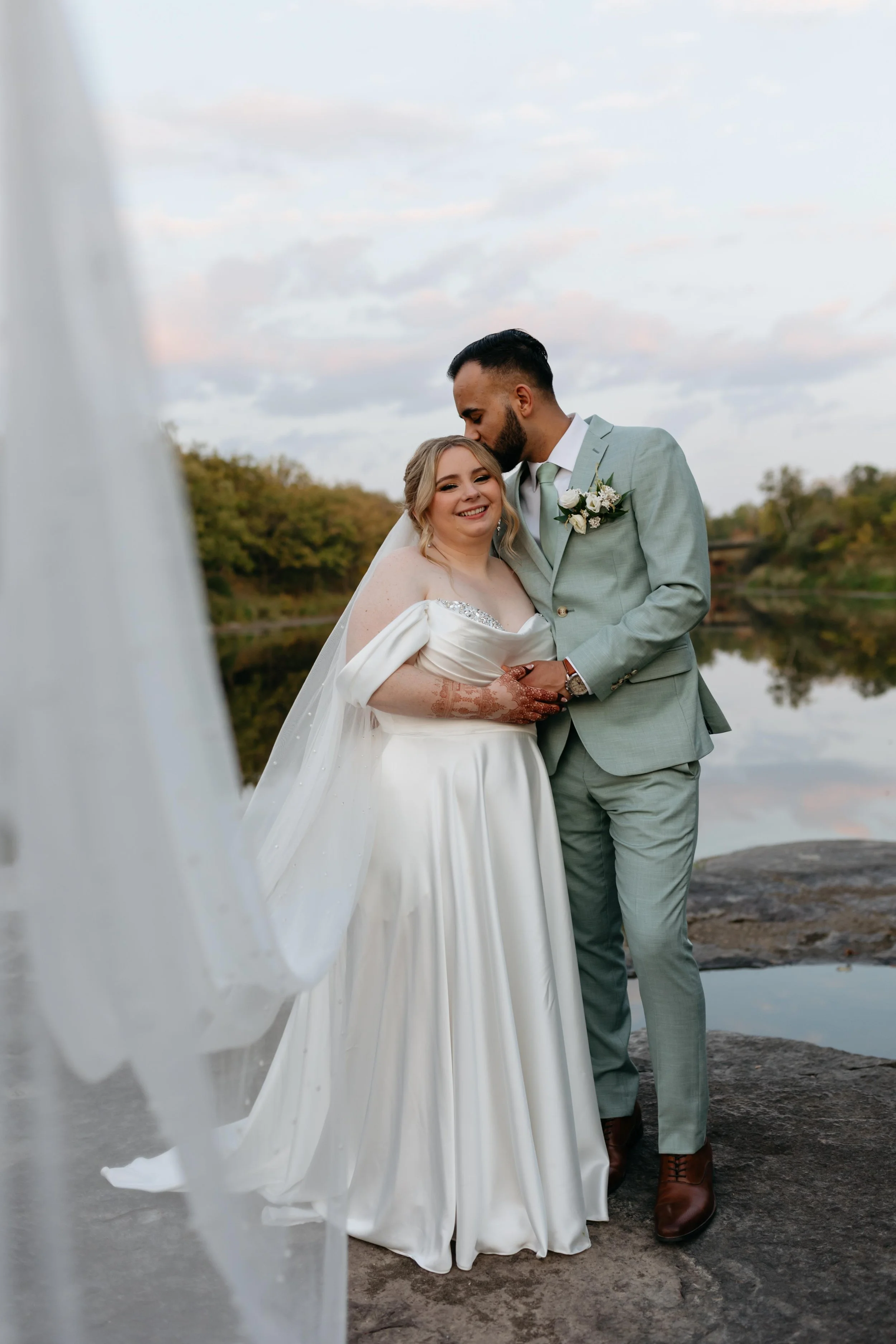 A bride and groom sharing an intimate moment outdoors near a body of water, with trees in the background under a partly cloudy sky. The groom is kissing the bride on her forehead, and they are both smiling. The bride is wearing a white wedding gown w