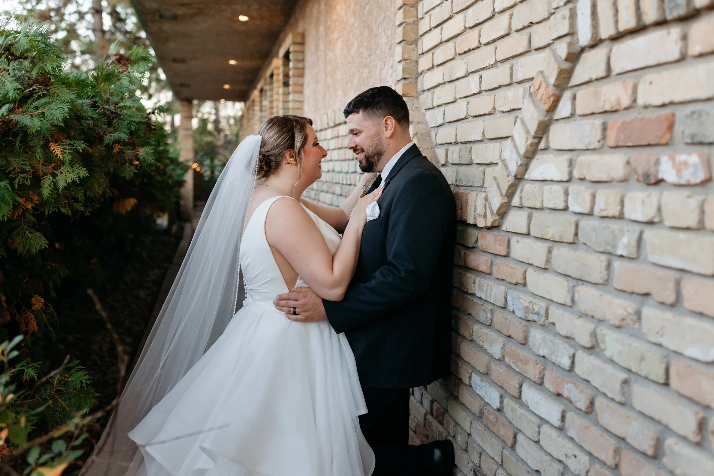 A bride and groom are smiling while posing close to each other beside a brick wall, with a garden on the left side. The bride is wearing a white wedding dress and veil, and the groom is dressed in a suit.