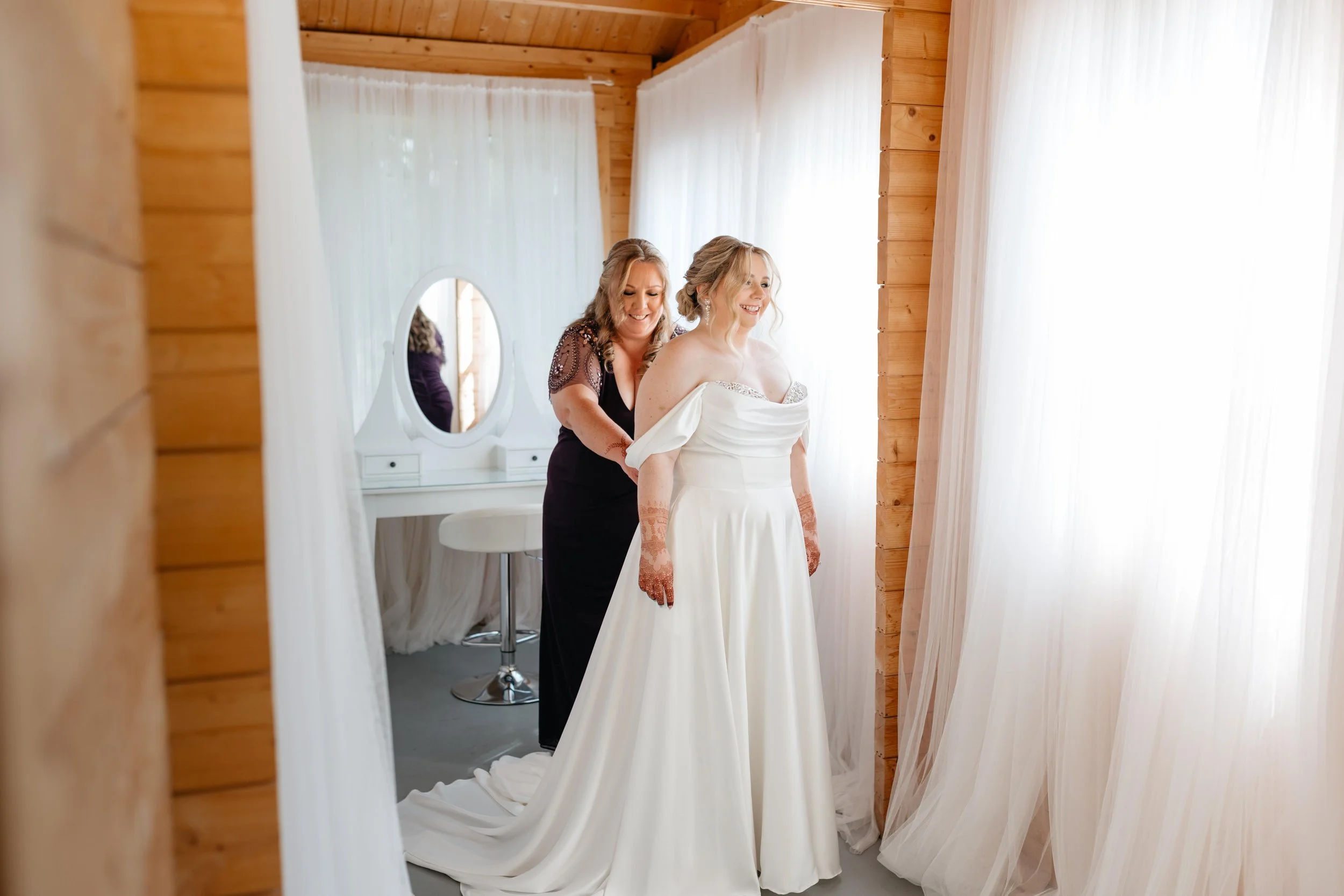 A bride getting ready in a room with wooden walls, assisted by a woman, while standing in front of a mirror with white curtains.