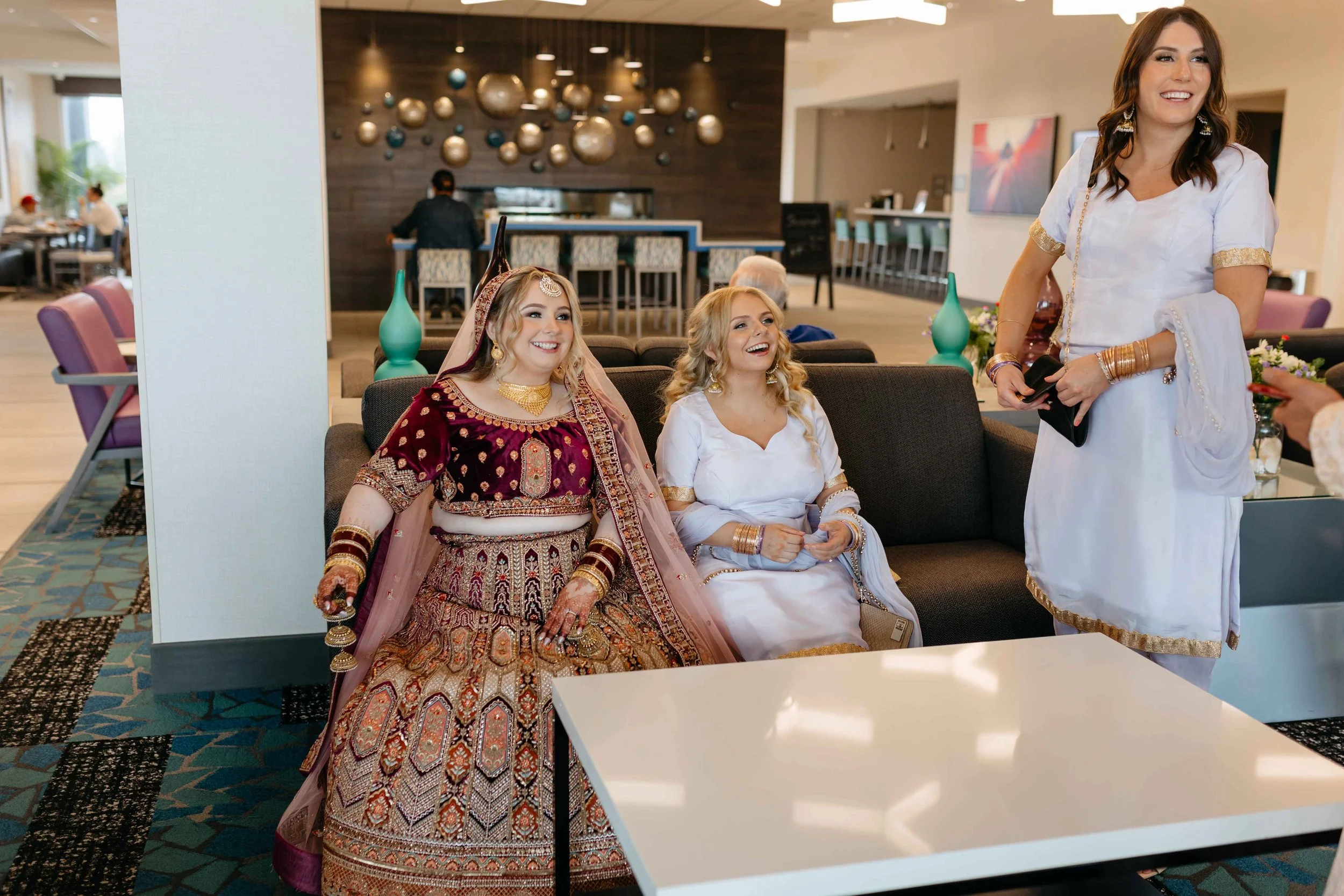 Women dressed in traditional South Asian attire seated on a couch, smiling, while another woman in a white dress stands nearby in a modern indoor setting with abstract wall art and bar area.