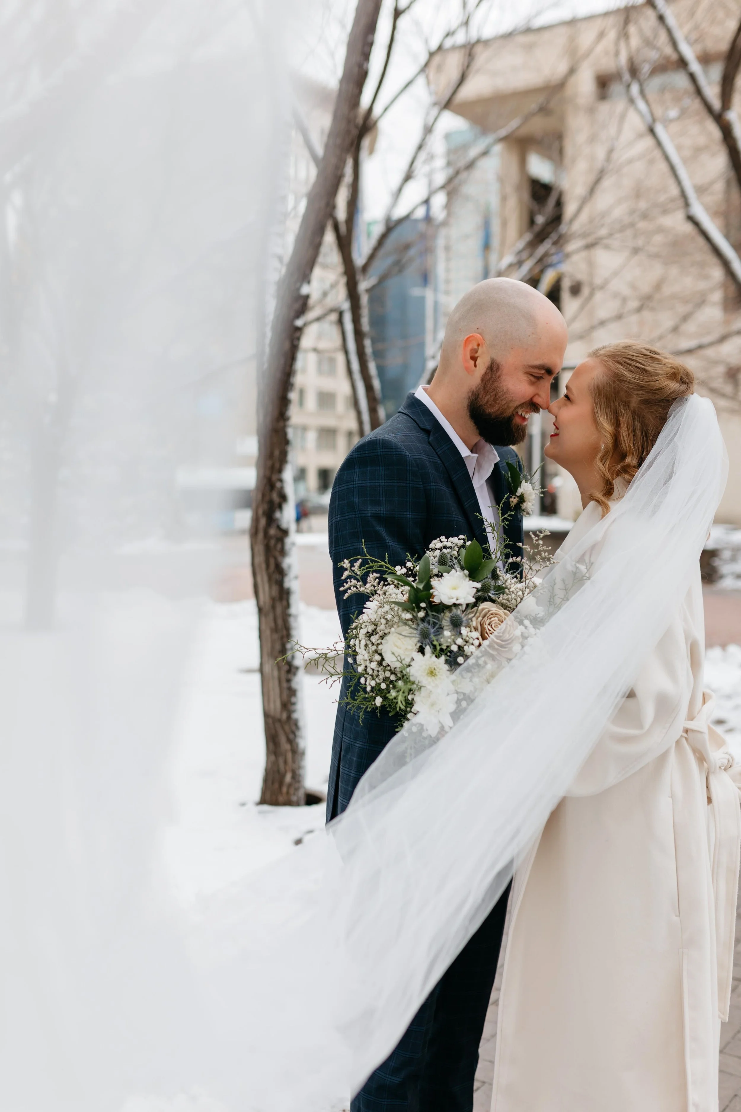 A couple on their wedding day, standing close with foreheads touching and smiling, outdoors in winter with snowy trees and urban buildings in the background.