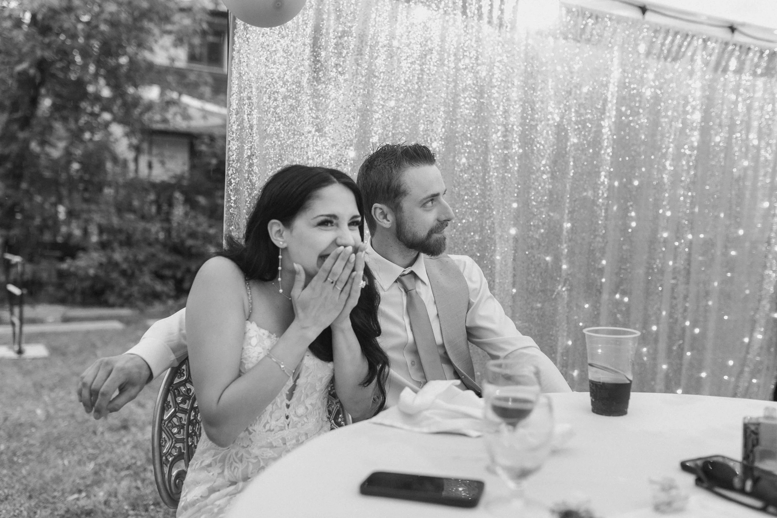 A black and white photo of a woman and a man sitting at a table outdoors, with the woman covering her mouth in surprise. The woman is wearing a lace dress and jewelry, and the man is dressed in a shirt with a vest. There is a glittering backdrop and 