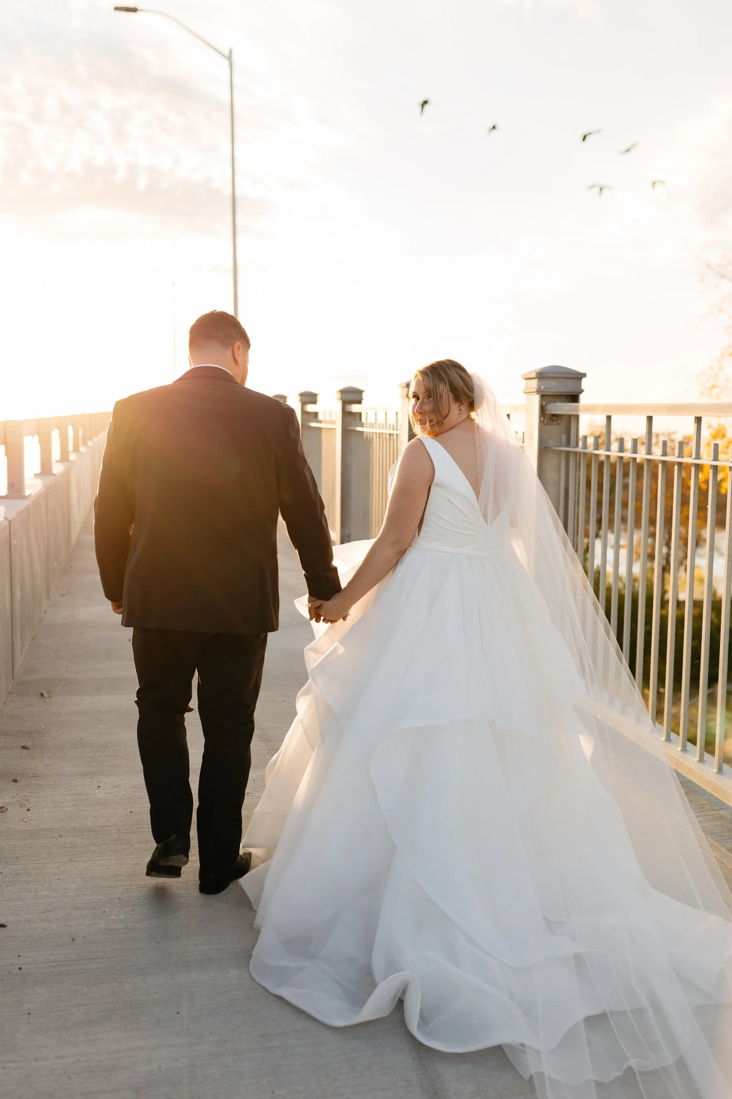 A newlywed couple walking hand in hand on a bridge at sunset, with the bride in a white wedding gown and the groom in a dark suit, looking back at each other and smiling, with birds flying in the sky above.