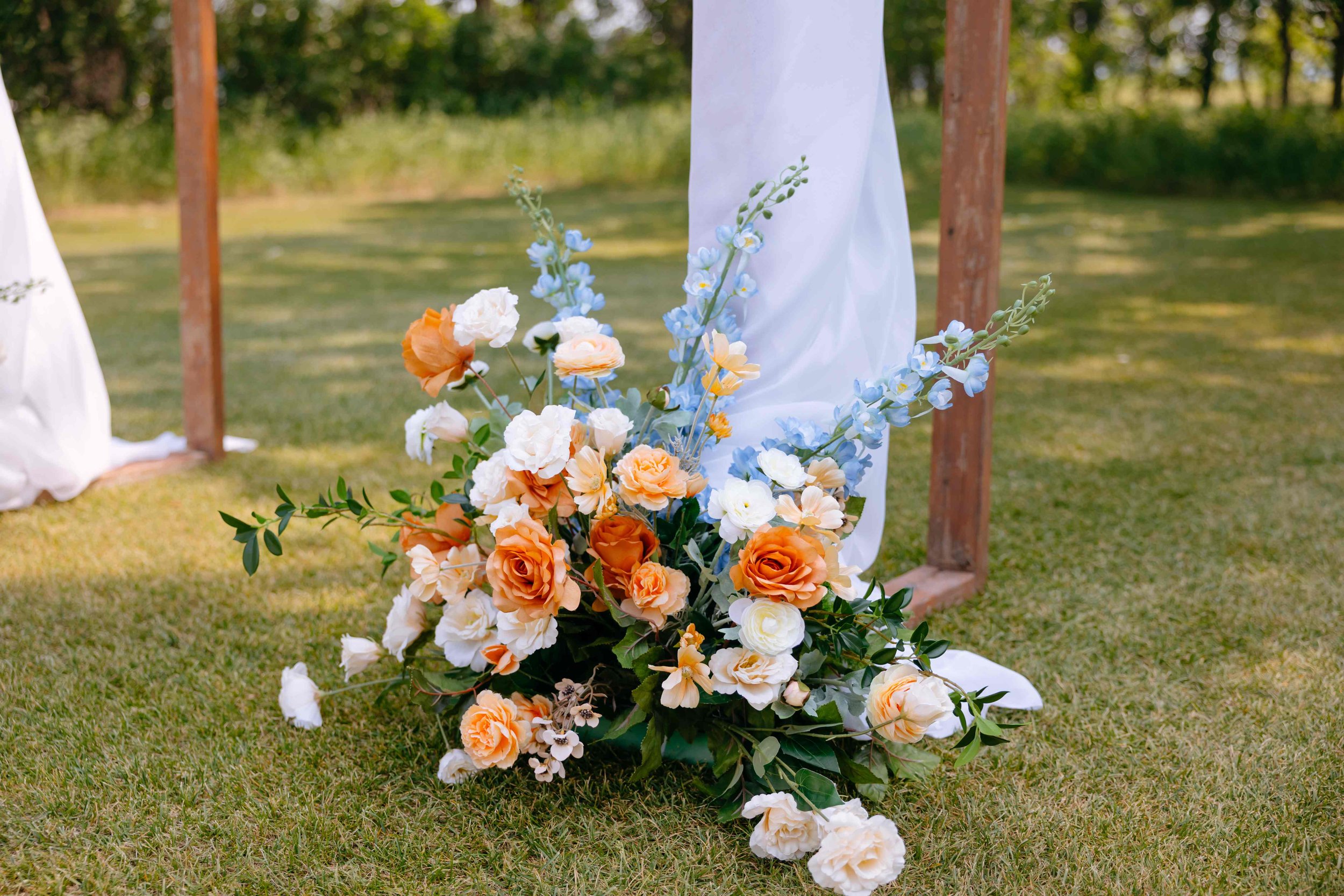 A floral arrangement featuring peach, white, and blue flowers on grass with a wooden wedding arch and draped white fabric in the background.