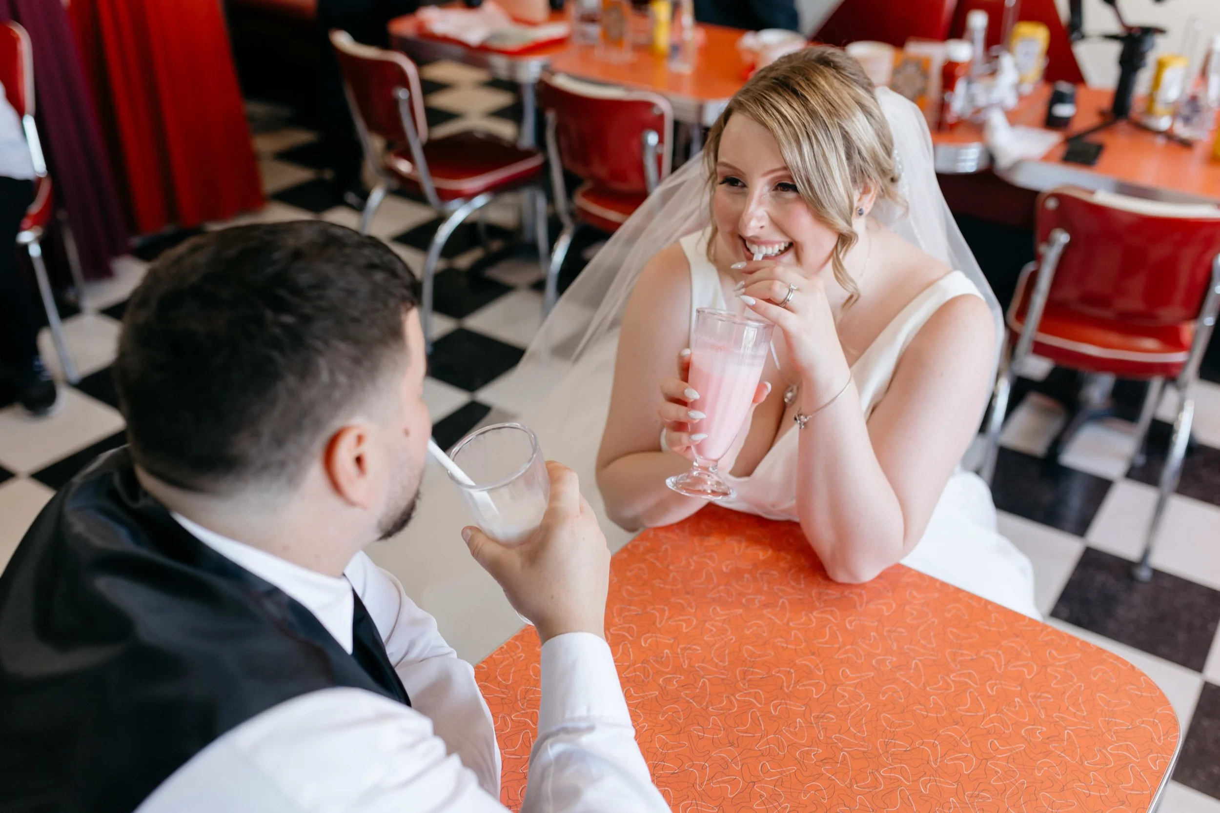 A bride with blonde hair in a wedding dress and veil smiling and talking to a groom in a tuxedo while holding a pink milkshake at a wedding reception.