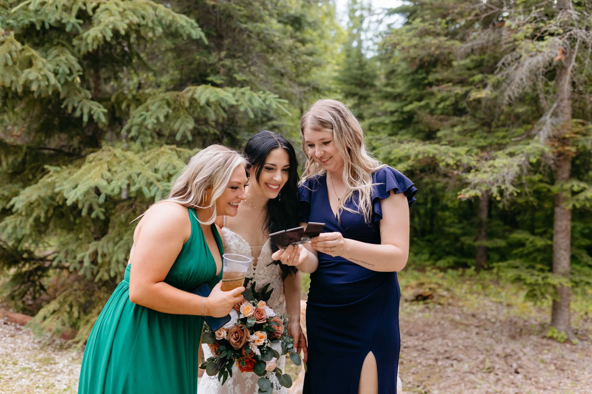Four women in formal dresses stand together outdoors, looking at something on a phone, with one holding a drink and a bouquet of flowers.