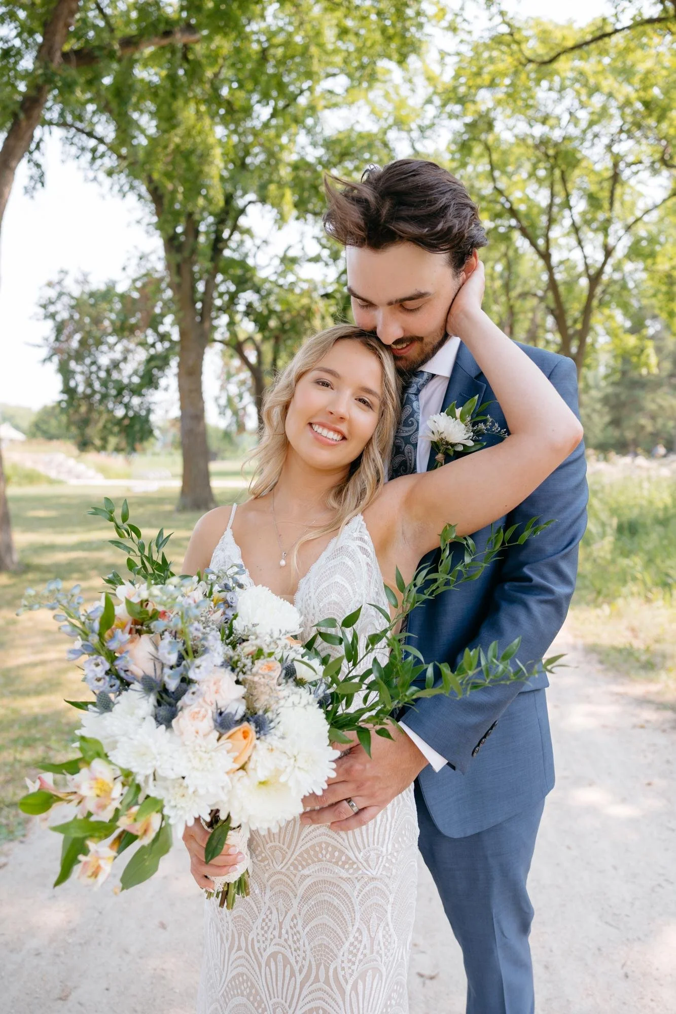 A newlywed couple with the bride holding a large bouquet of flowers, standing outdoors near trees. The bride is smiling with her eyes closed, wearing a white lace wedding dress. The groom, dressed in a blue suit, is leaning his forehead gently against her head, with her hand on his face. The scene is bright and cheerful in a park-like setting.