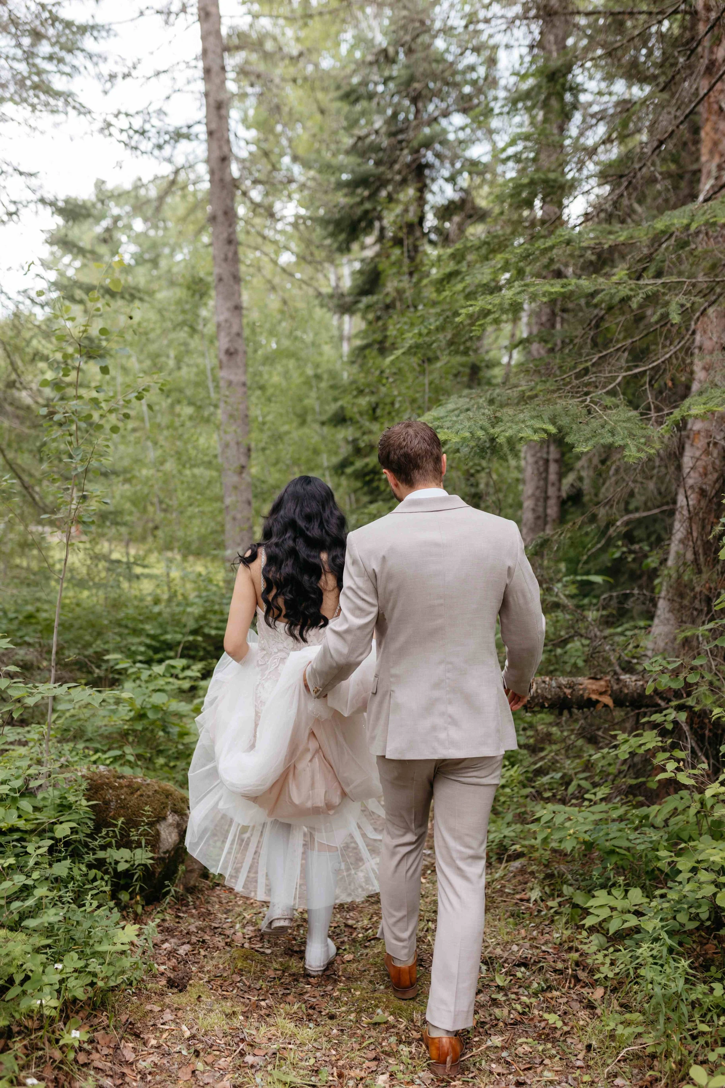 A couple dressed in wedding attire walking through a wooded forest trail.
