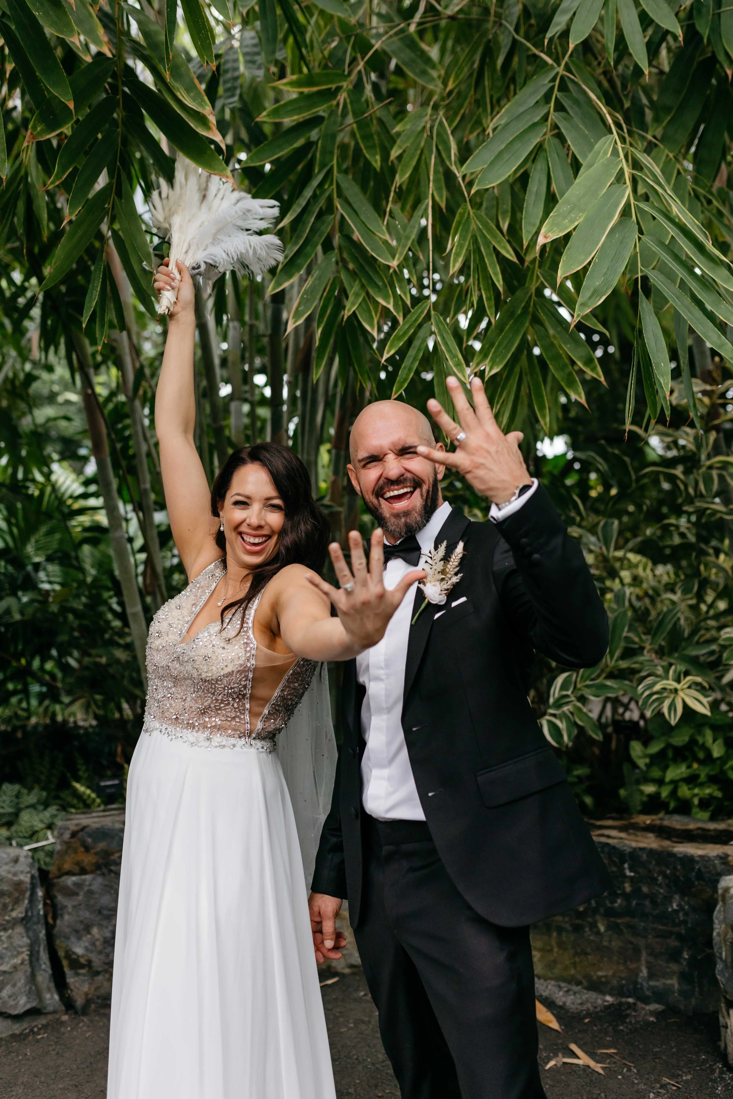 A bride and groom celebrate outdoors among green leafy trees, smiling and making playful hand gestures. The bride holds a white feather fan, and the groom wears a black tuxedo with a boutonniere.