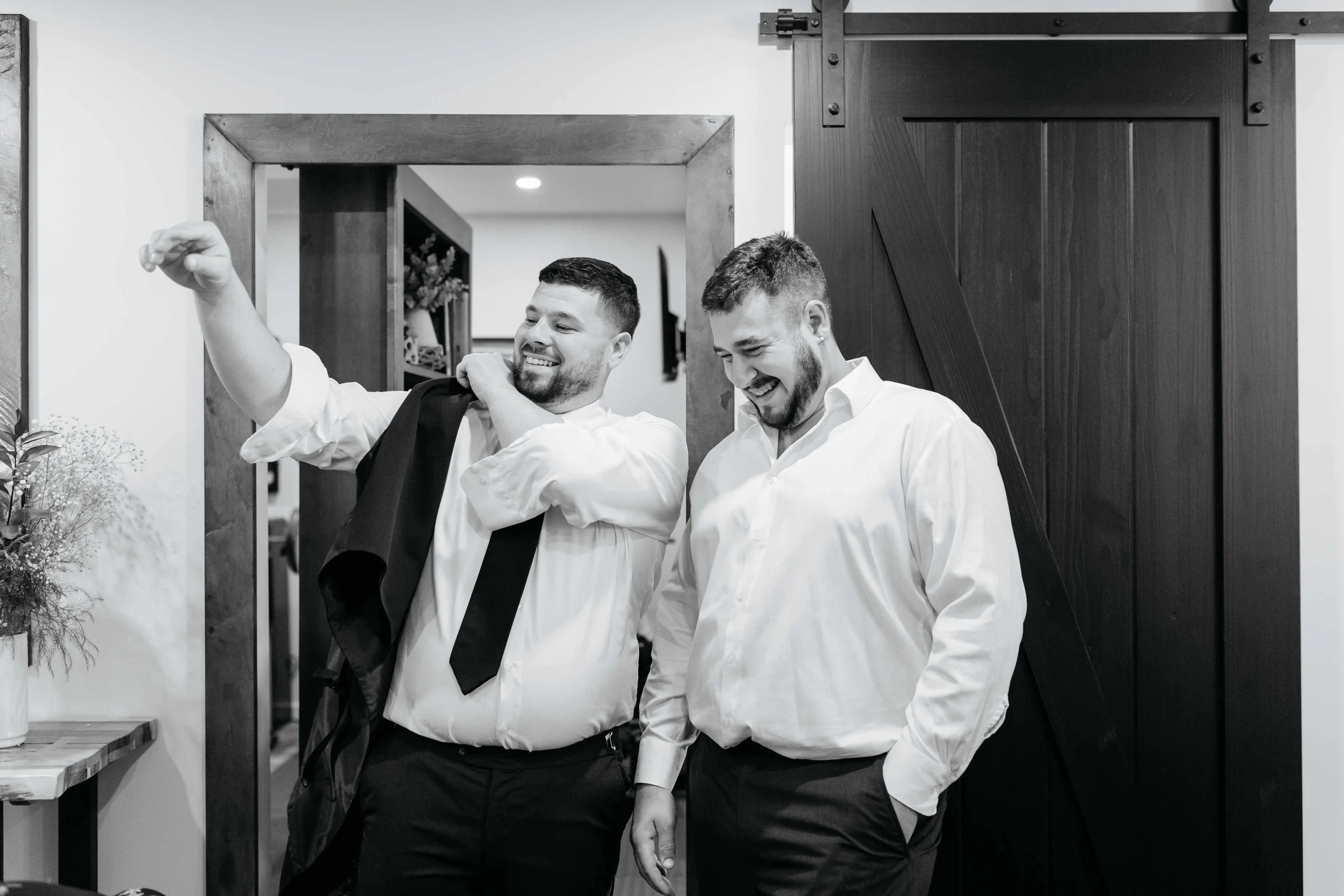 Two smiling men dressed in white shirts, one with a jacket over his shoulder, standing near a wooden sliding door inside a home or office, engaged in a moment of laughter.