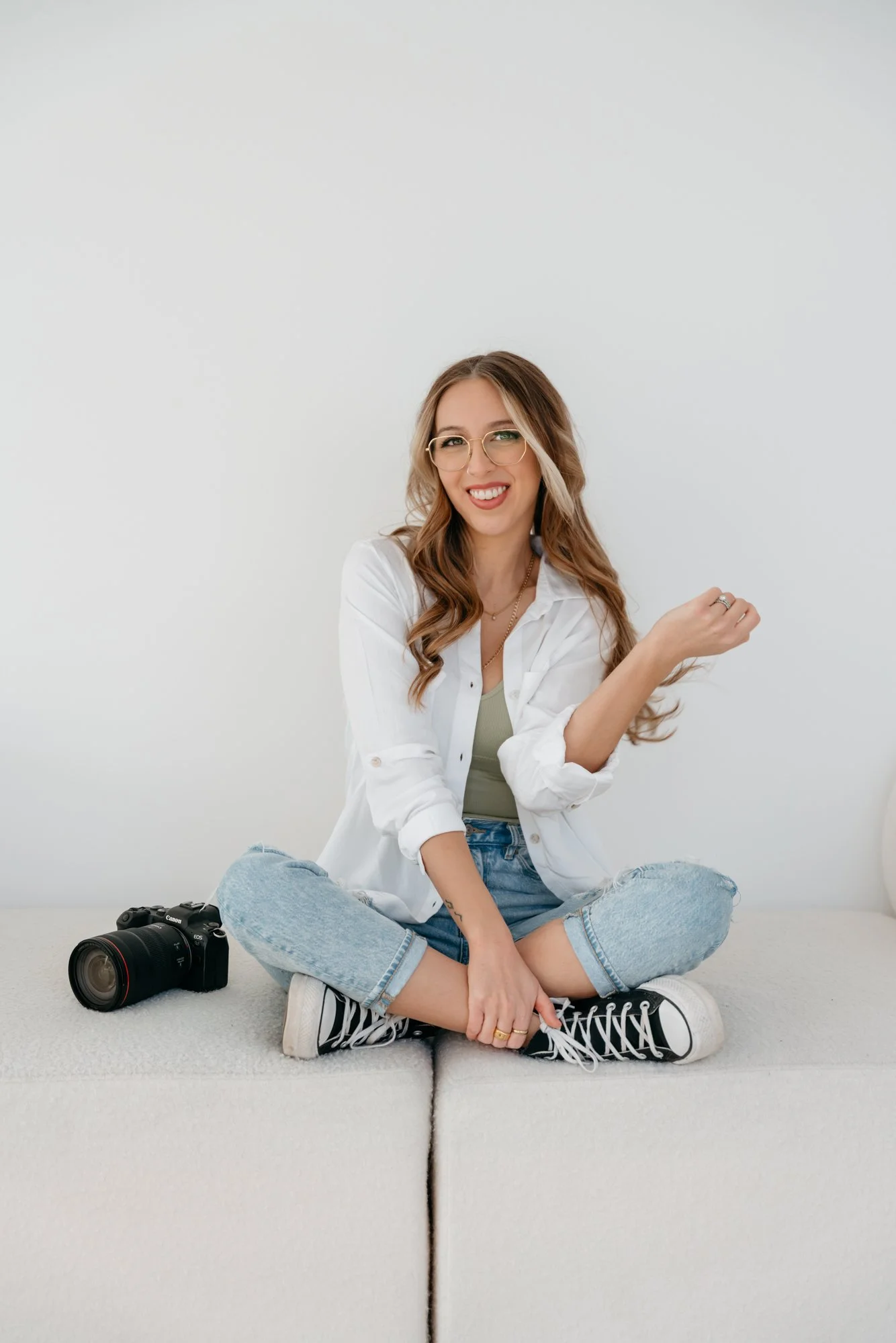 Sari Riseby, a Winnipeg family and newborn photographer, is sitting cross-legged in a white shirt and jeans, smiling and looking at the camera, with a DSLR camera on the floor nearby.