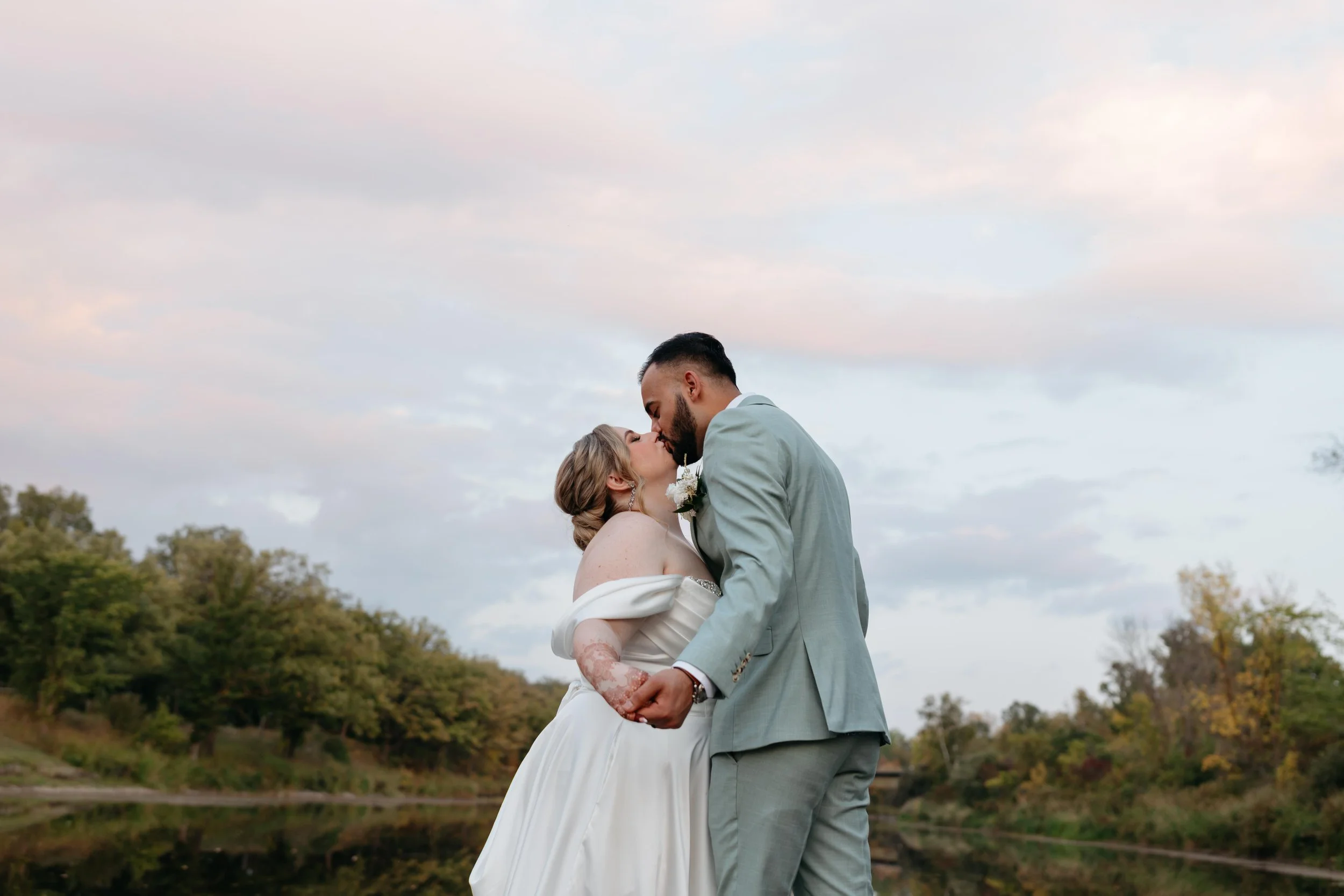 A bride and groom share a kiss outdoors during their wedding, with trees and a cloudy sky in the background.