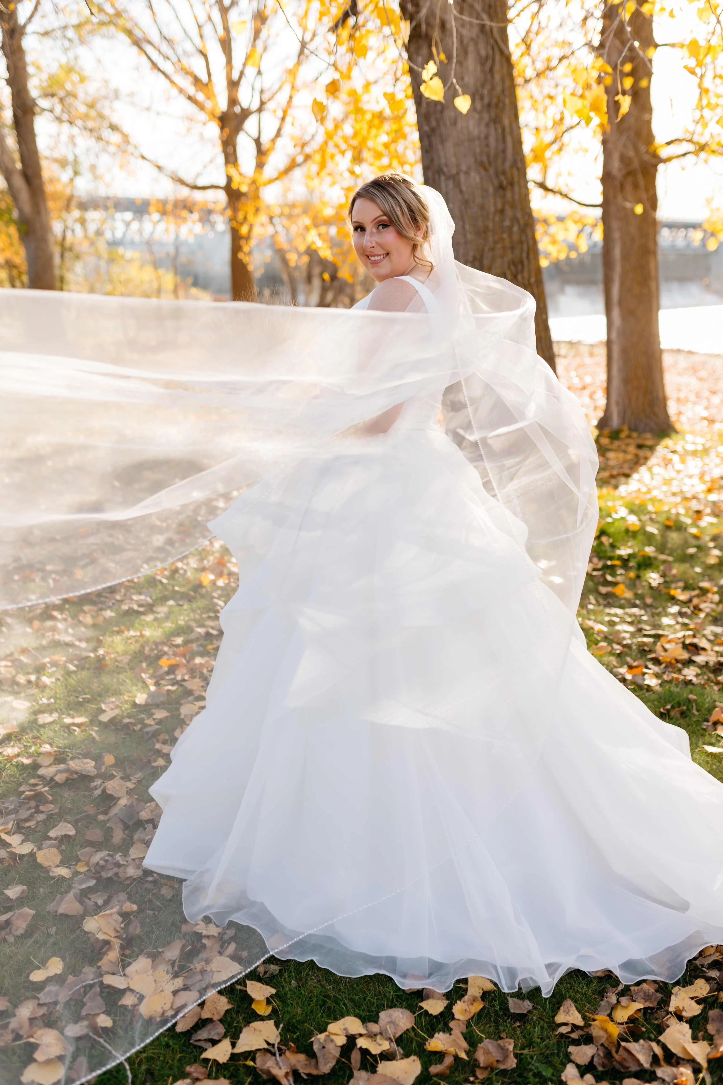 A bride in a white wedding gown with a long flowing veil outdoors in autumn, surrounded by trees with yellow leaves and fallen leaves on the ground, smiling and looking back over her shoulder.