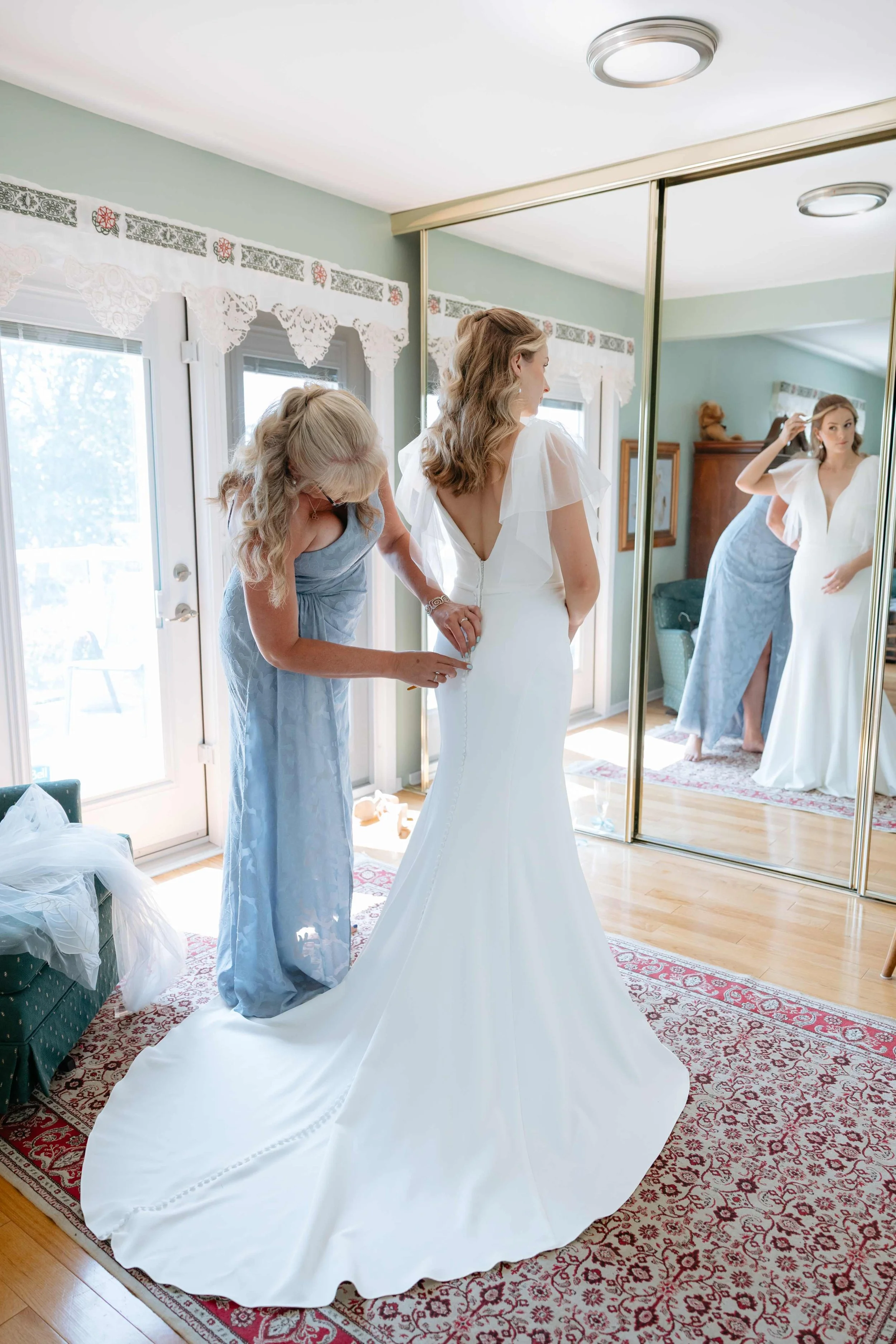 A bride in a white wedding dress standing in front of a mirror, helping her by a woman in a light blue gown inside a room with a carpet, large window, and mirrored closet doors.