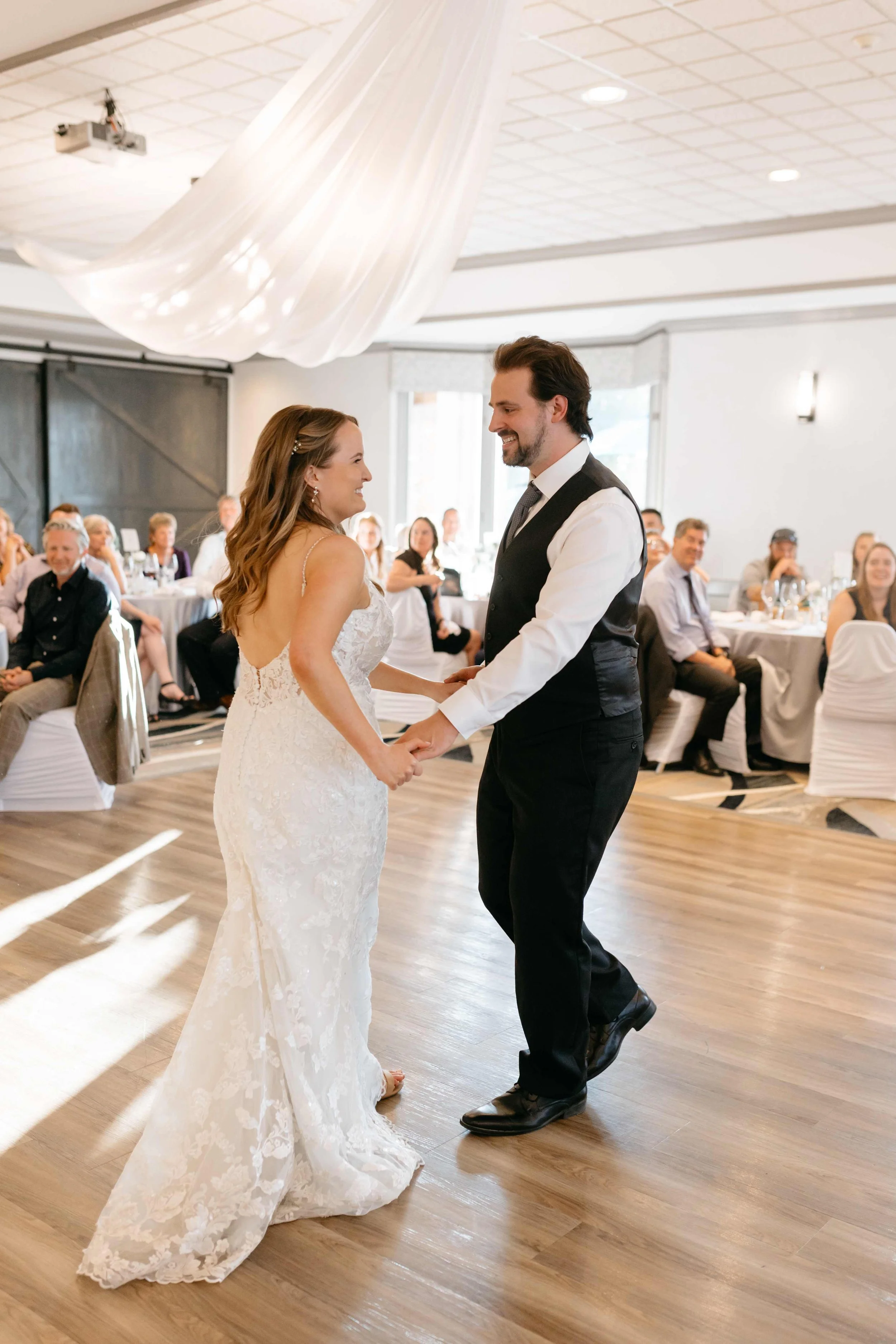 A bride and groom are holding hands and dancing during their wedding reception in a banquet hall filled with seated guests. The bride wears a white lace wedding gown, and the groom wears a black shirt, black vest, and black pants. The hall has a wood