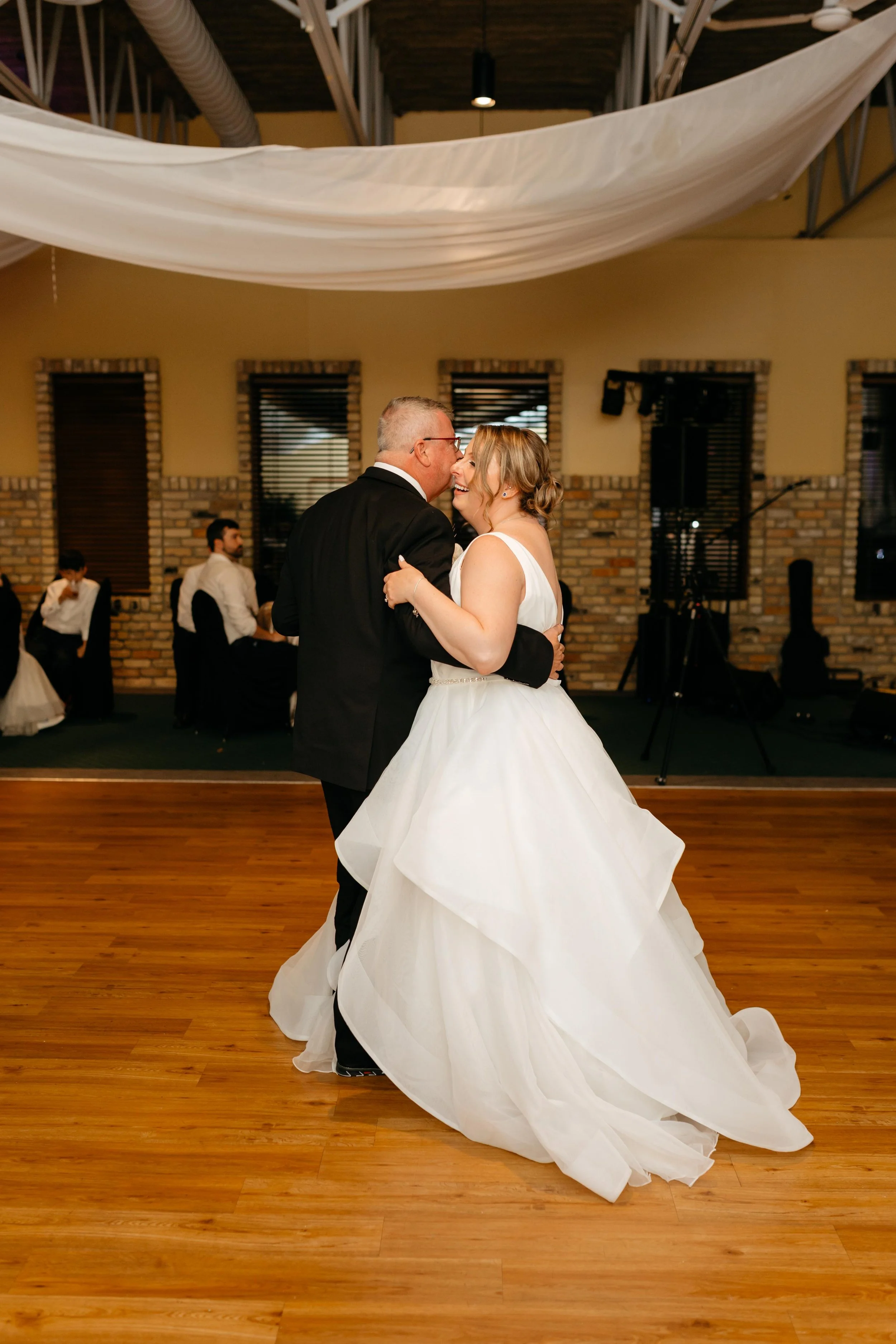 A bride and groom sharing a dance at their wedding reception in a rustic venue with wood floors, brick walls, and large windows, decorated with a white draped fabric ceiling.