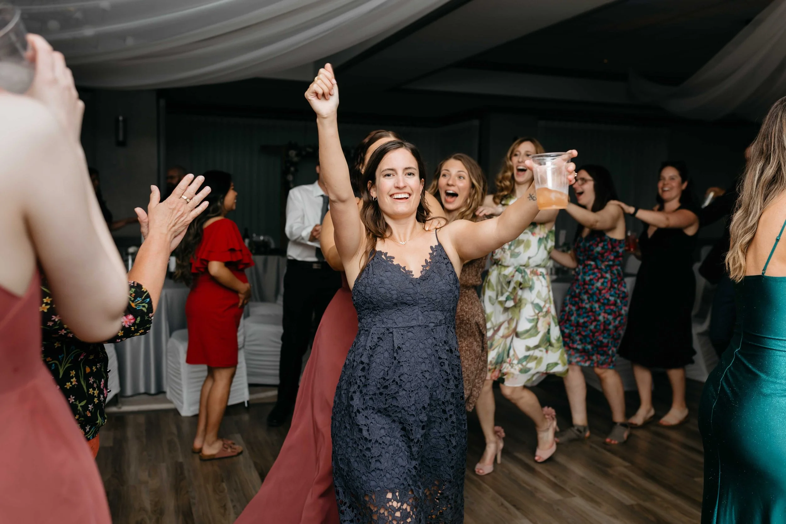 A group of women dancing and celebrating at a party, with one woman in a blue dress raising her hand and holding a drink.