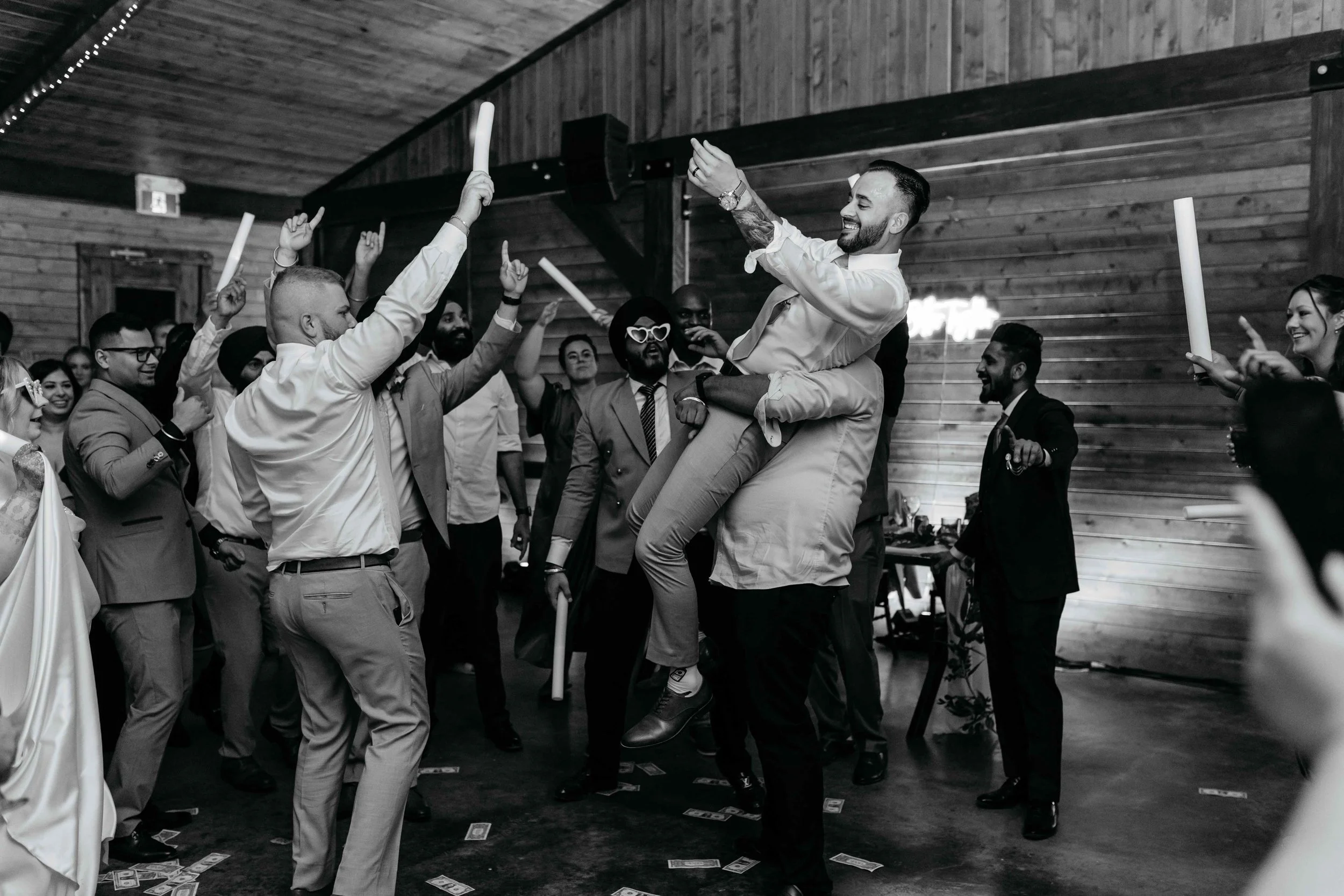 Group of people celebrating at a party, with a man being lifted and others holding foam sticks, in a room with wooden walls.