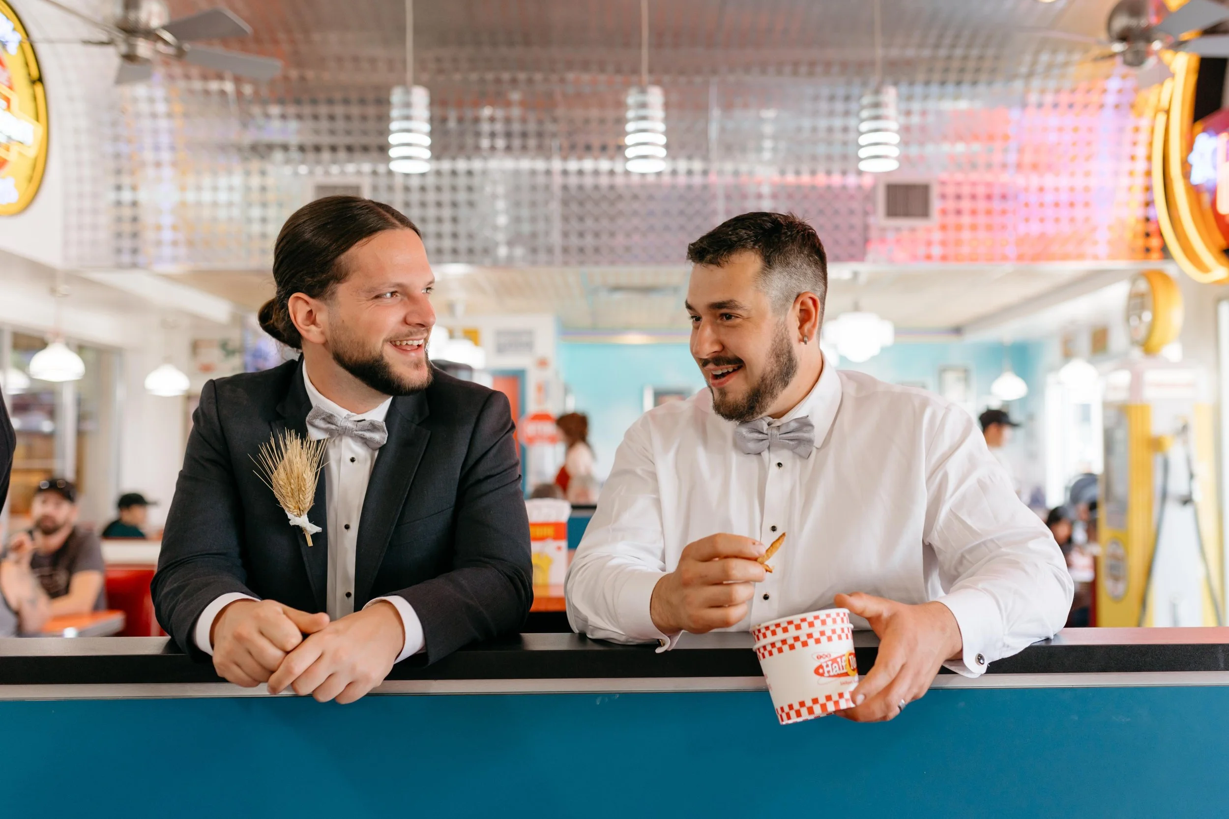 Two men in tuxedos with bow ties sitting at a counter in a retro-style diner, smiling and talking, one holding a cup of soft serve ice cream.