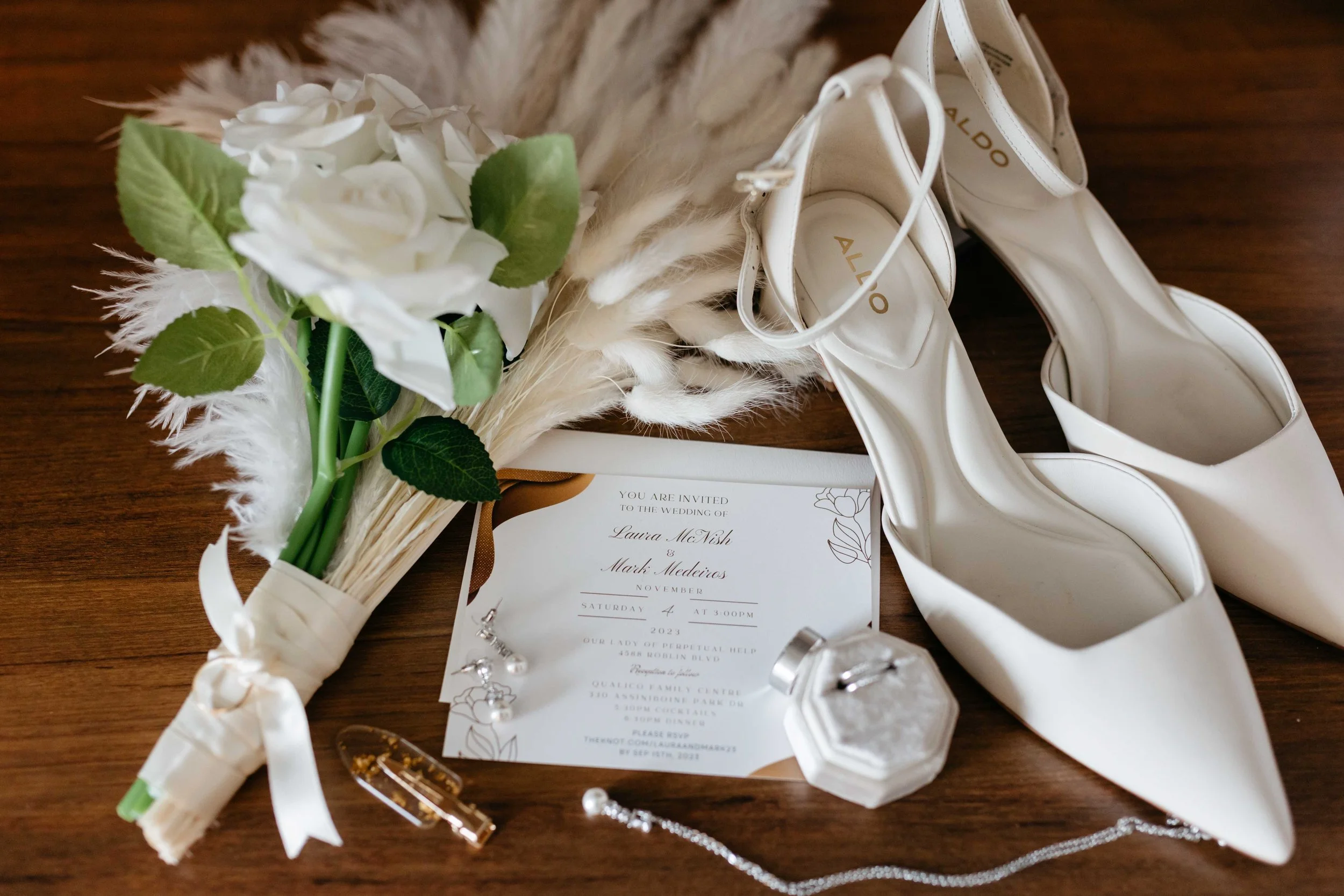 A wedding invitation, white high-heeled shoes, a small bouquet of white flowers with green leaves, and jewelry including rings and earrings are displayed on a wooden surface.