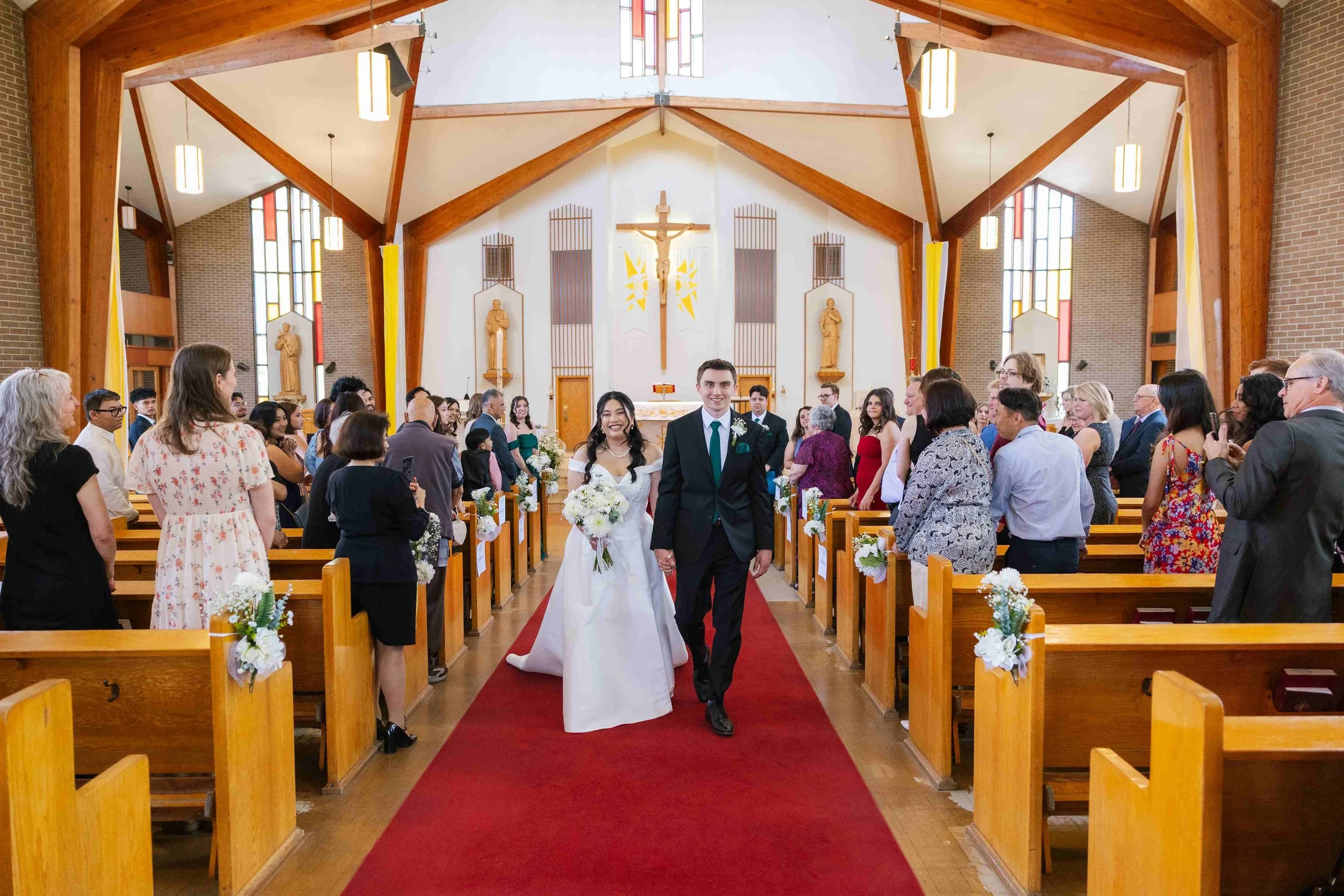 A bride and groom walking down the aisle afterward of their wedding ceremony in a church filled with guests on either side of the aisle, with the altar and religious statues in the background.