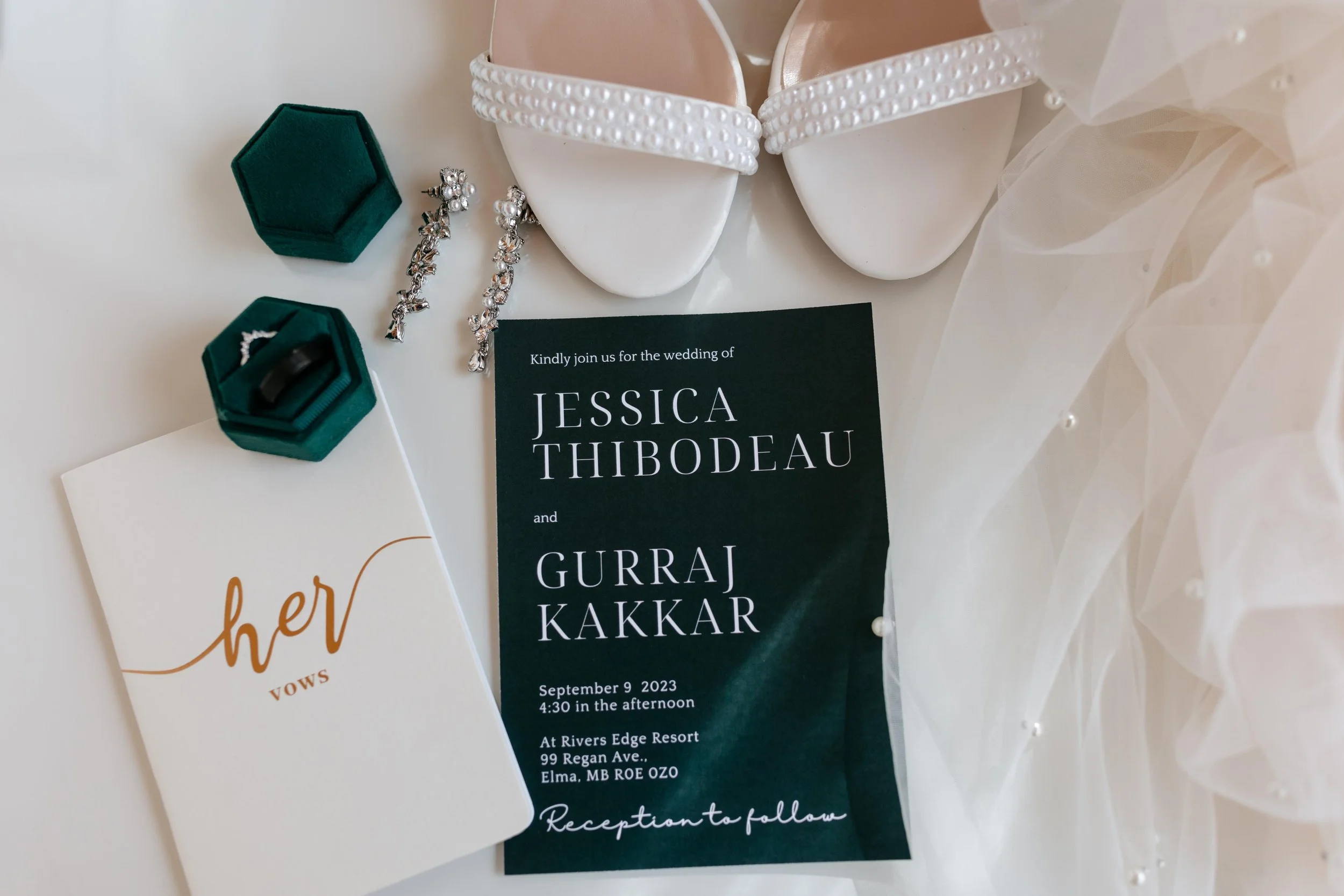 White wedding shoes with pearl details, silver earrings with pearls, a black wedding invitation with white text, green rings box with rings, and a wedding invitation card.