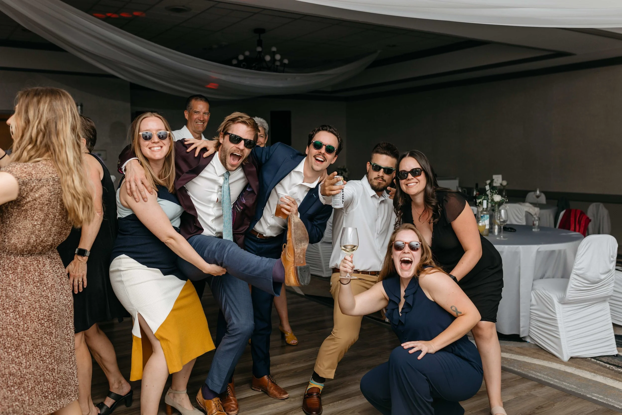 A group of people at a celebration or party, smiling and having fun. Some are wearing sunglasses and holding drinks, with a decorated event space in the background.