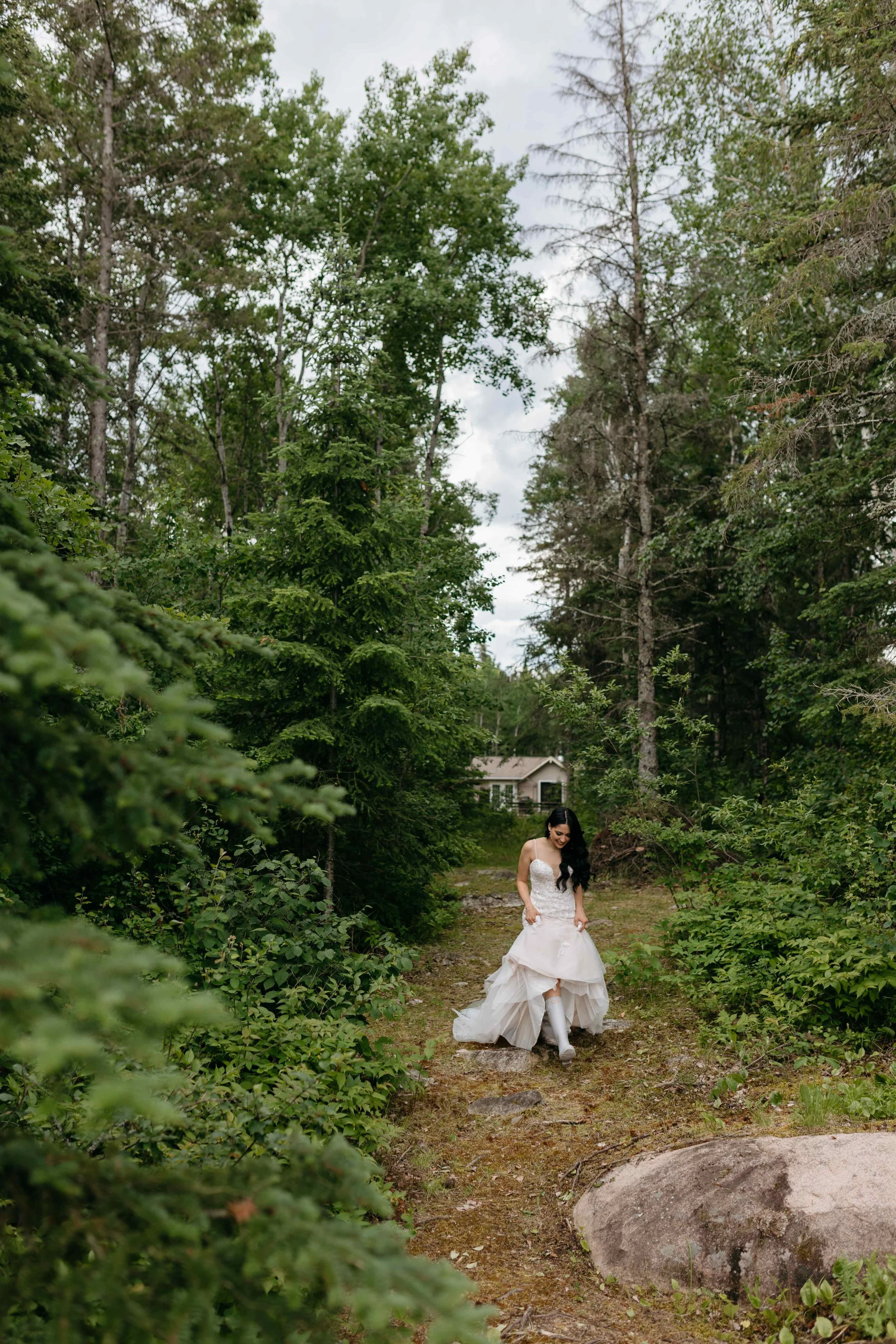 A woman in a white wedding dress walking on a dirt path in a lush green forest with trees on either side and a house visible in the background.