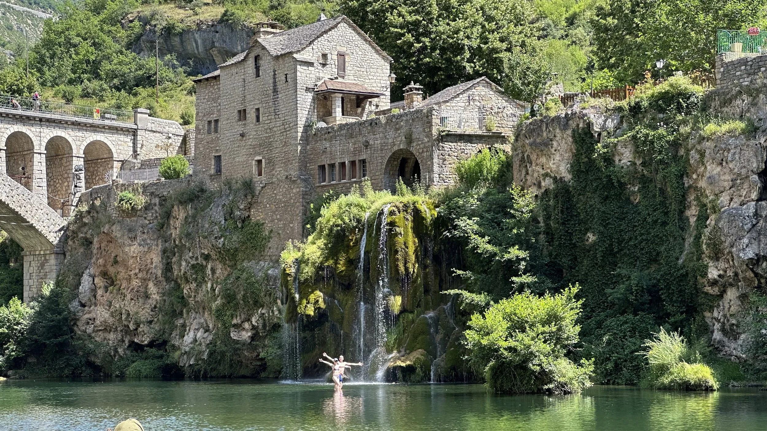 A person stands with arms outstretched at the edge of a small waterfall in a scenic area with stone buildings, bridges, lush greenery, and a body of water.