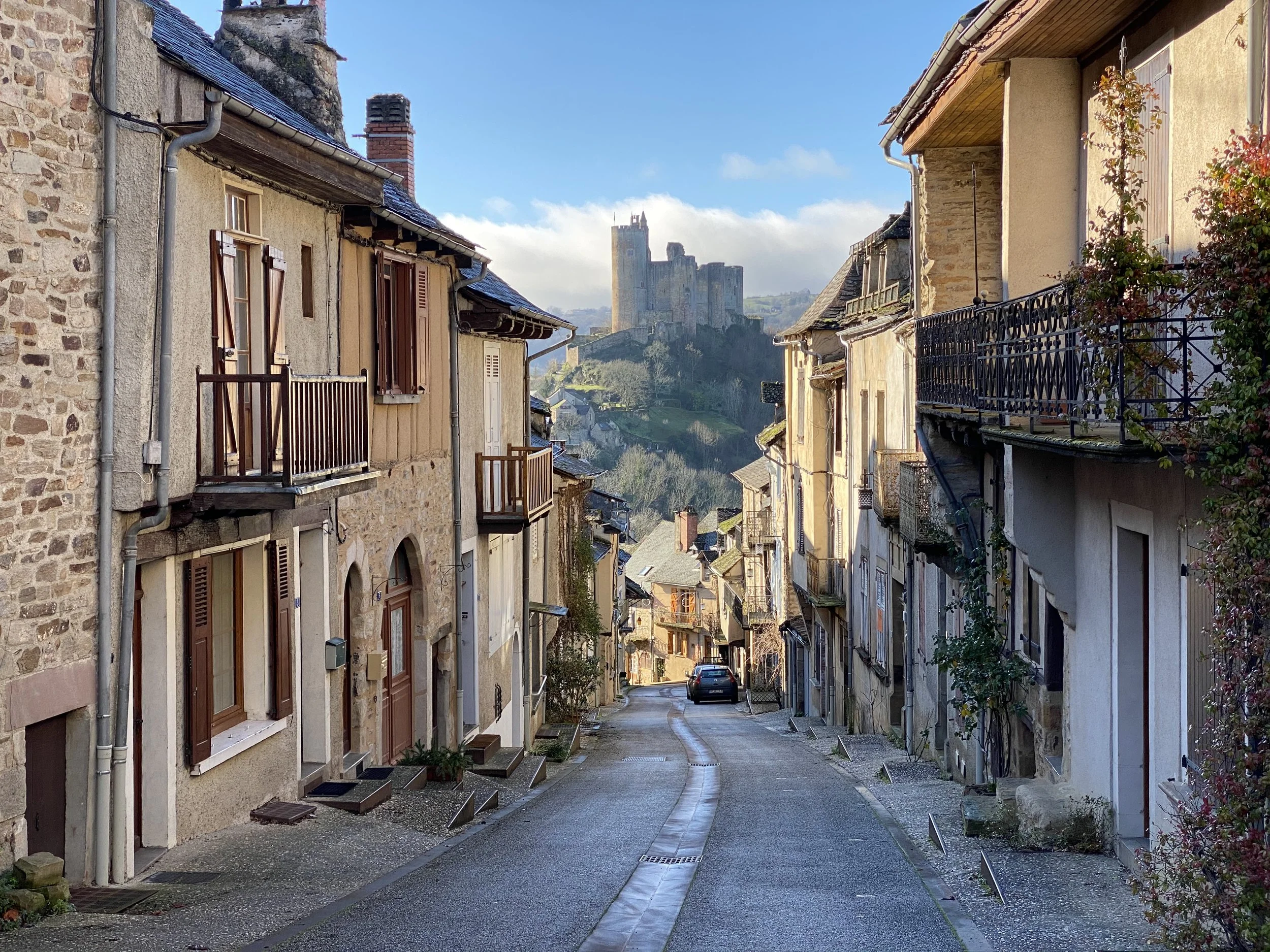 A narrow, winding cobblestone street lined with old stone and stucco houses with wooden shutters, leading to a distant hilltop castle in the background under a partly cloudy blue sky.