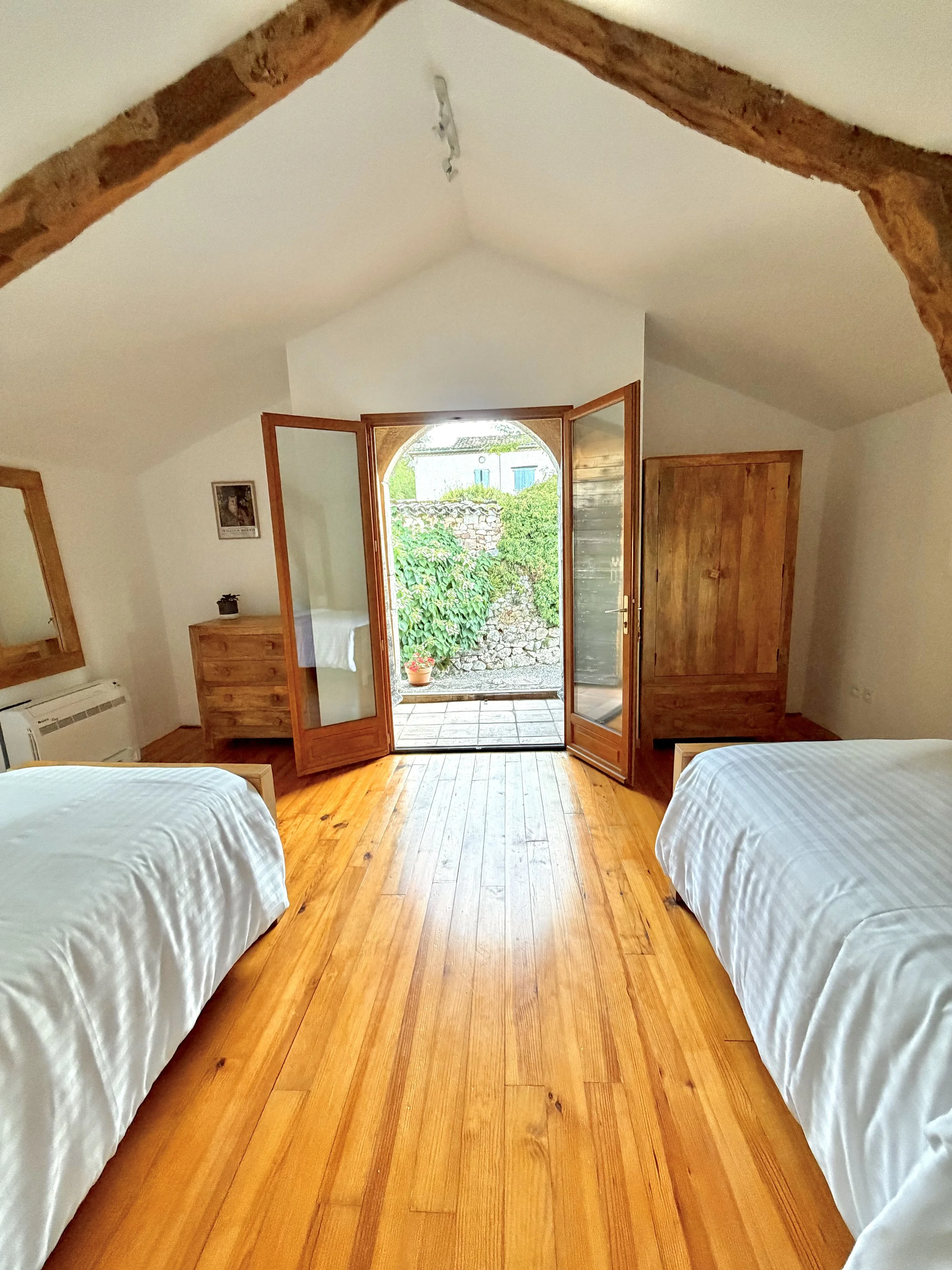 View of a bedroom with two beds covered in white linens, wooden furniture, a door opening to a patio with a stone wall and greenery outside, a small framed picture on the wall, and warm wooden flooring.