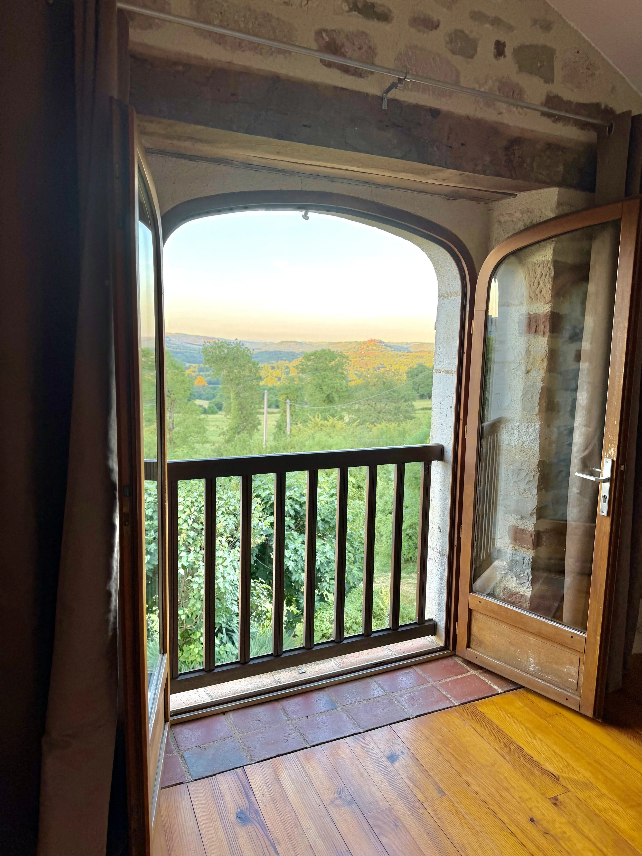 Open wooden window with a small balcony, showing green trees and rolling hills in the distance, under a light blue sky.