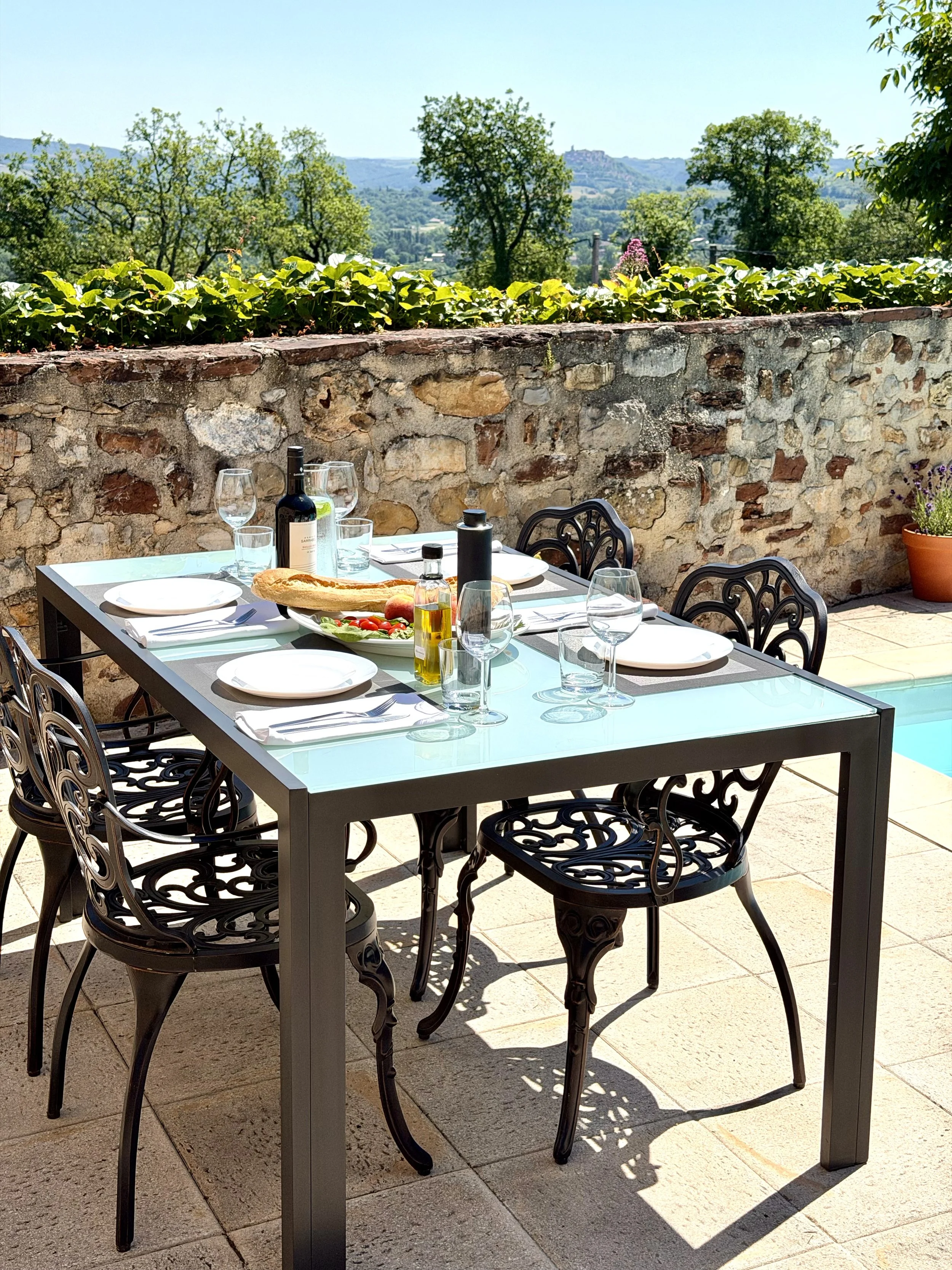 An outdoor dining table set with plates, glasses, a bottle of wine, olive oil, and bread, surrounded by ornate black chairs, with a stone wall, green plants, and scenic hills in the background.